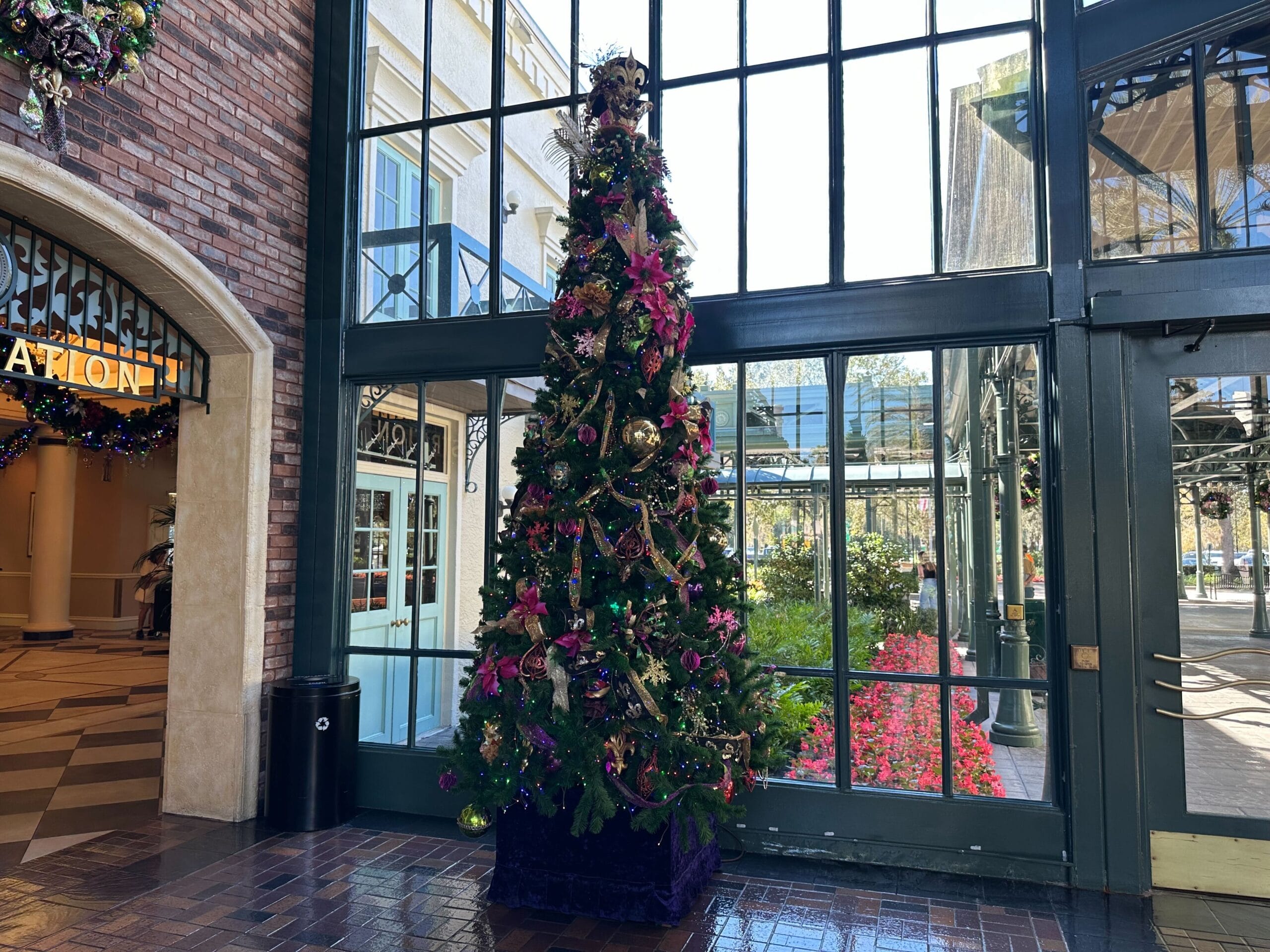 Tall Christmas tree with colorful ornaments stands in a brick and glass atrium. Sunlight filters through large windows, illuminating flowers outside.