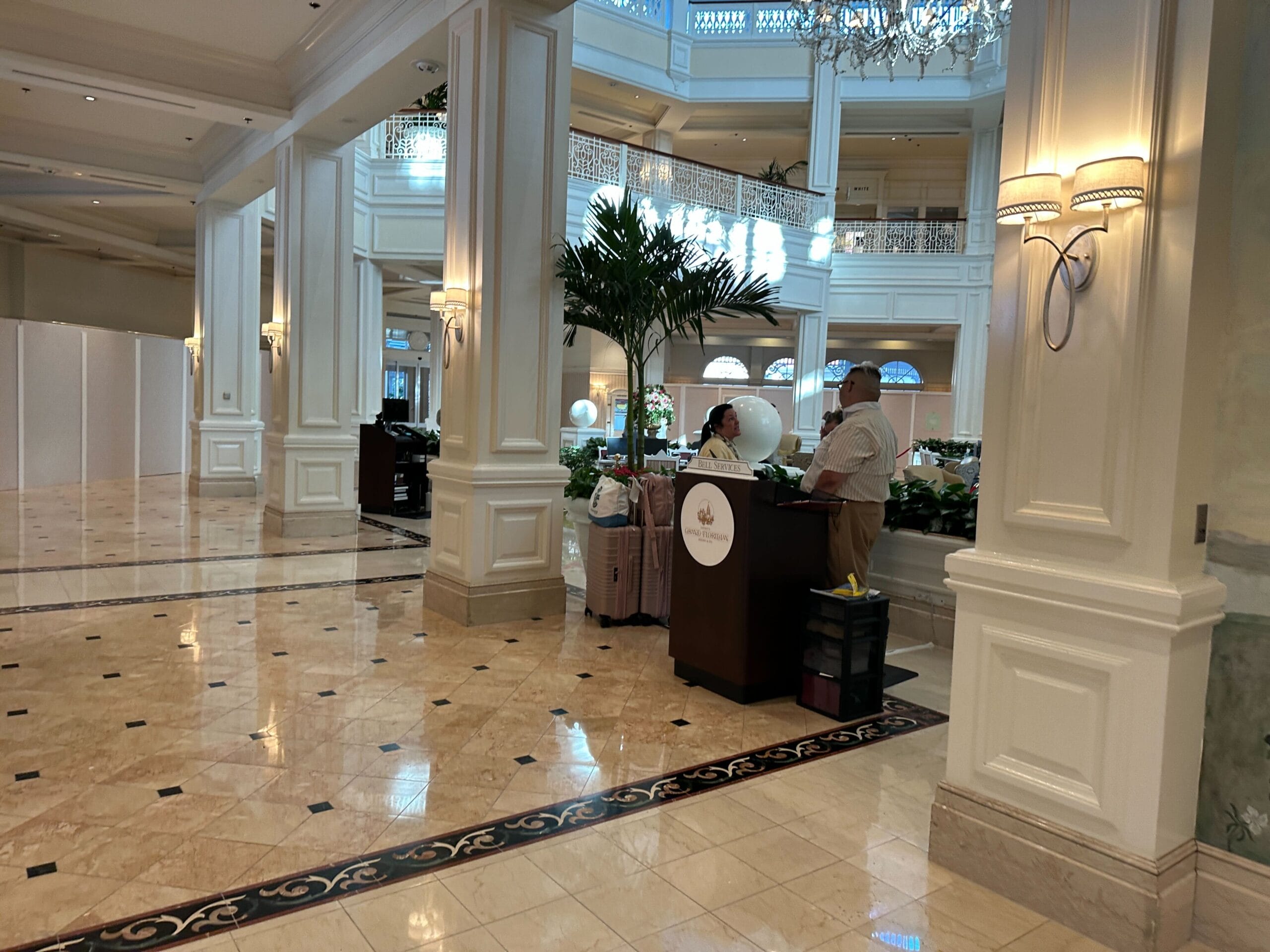 Two hotel staff stand behind a podium in a grand, marble-floored hotel lobby with columns and a potted palm.