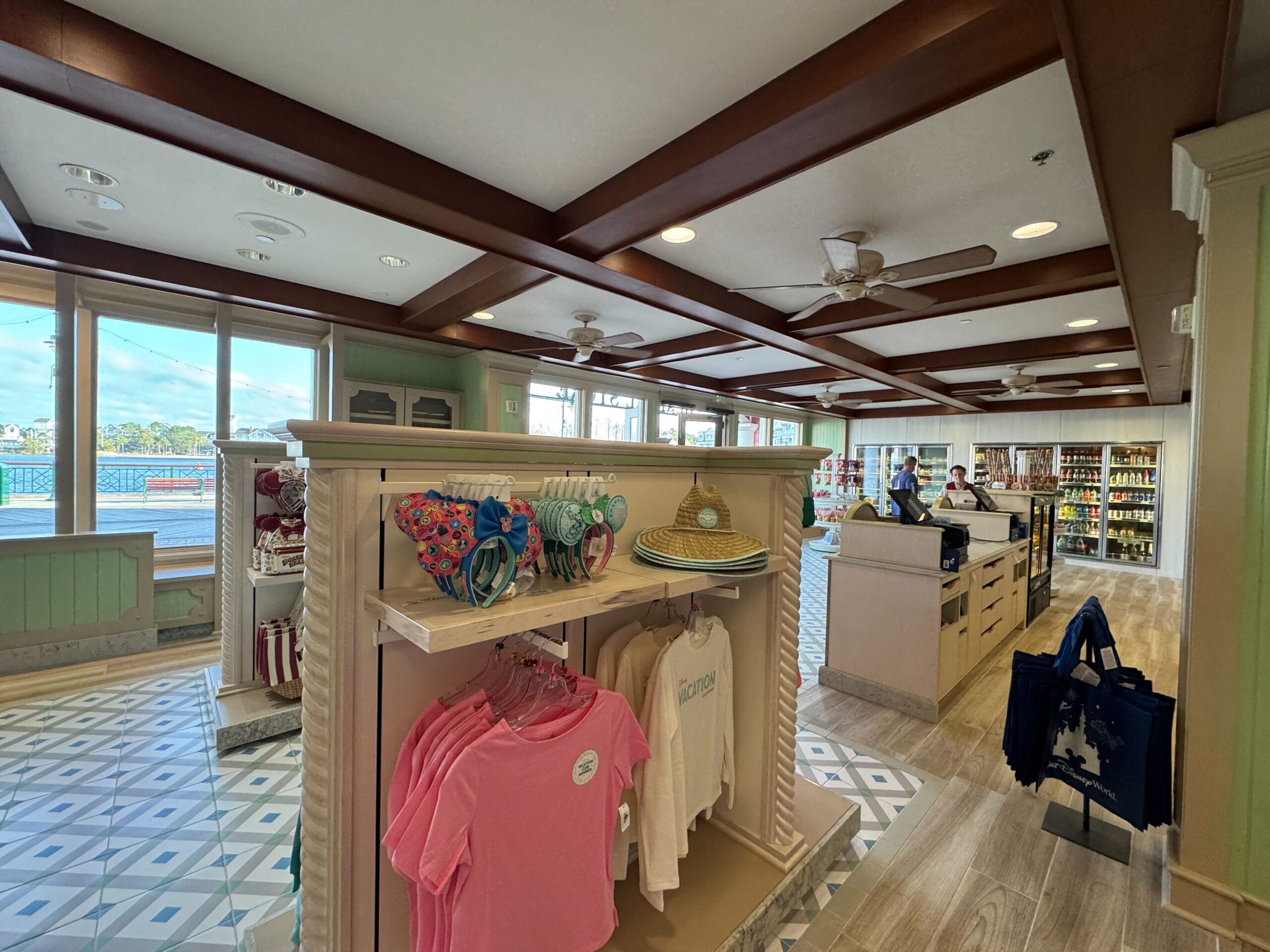Store interior with clothing and accessories displayed on racks, two people at the counter, and large windows showing an outside view.