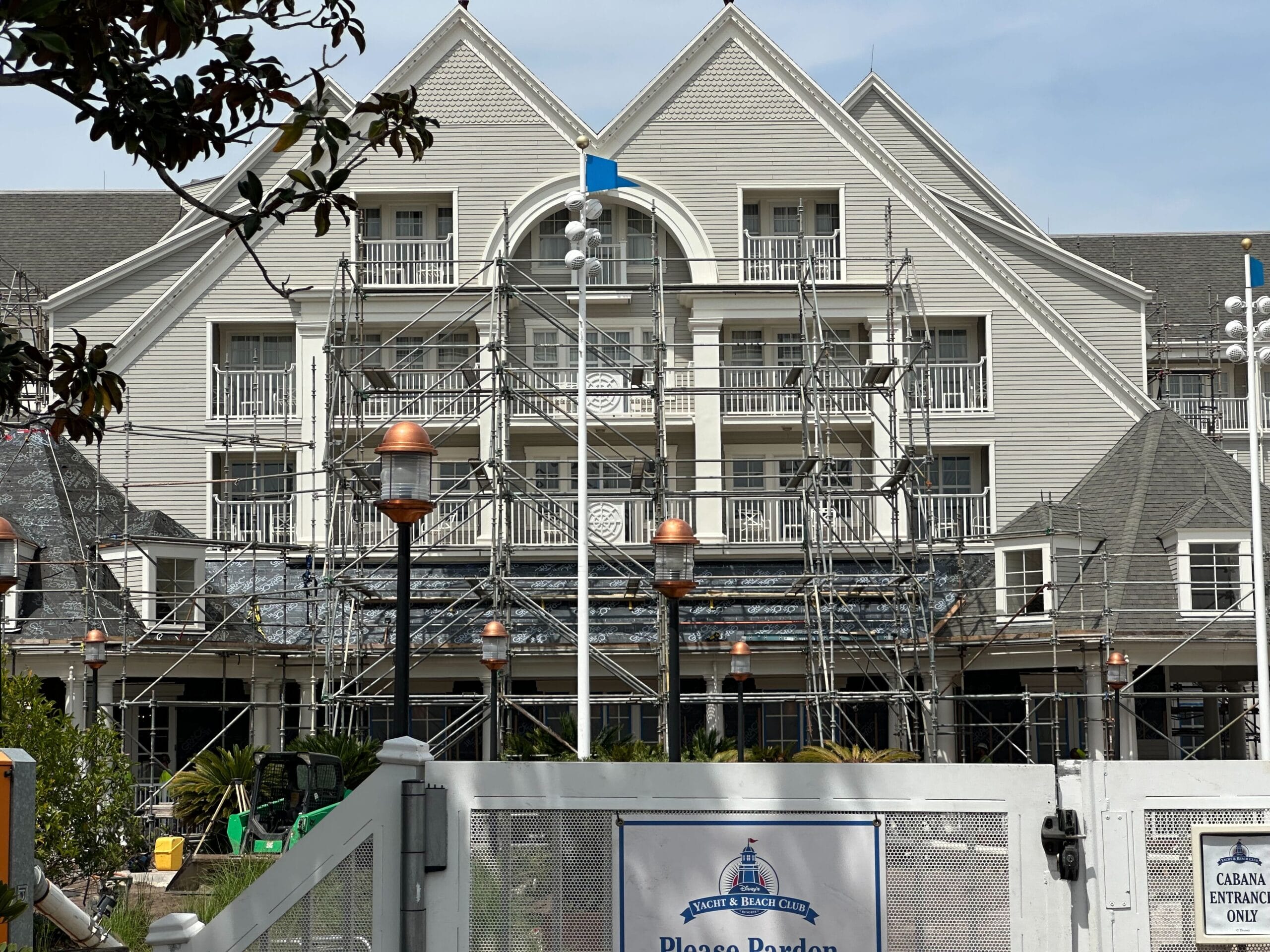 A large building undergoing maintenance, with scaffolding in place, resembles Disney's Yacht Club. The structure boasts a gray facade with white balconies and a steep roof. A sign reads Please Pardon Our Appearance as pool construction progresses.