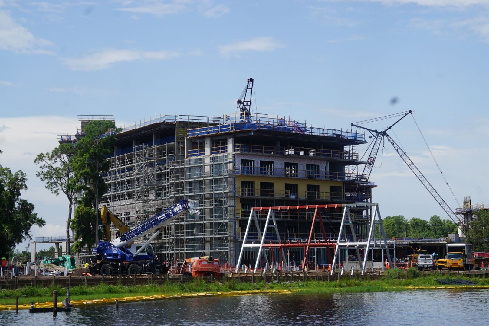 A multi-story building under construction with cranes and scaffolding by a body of water on a partly cloudy day.
