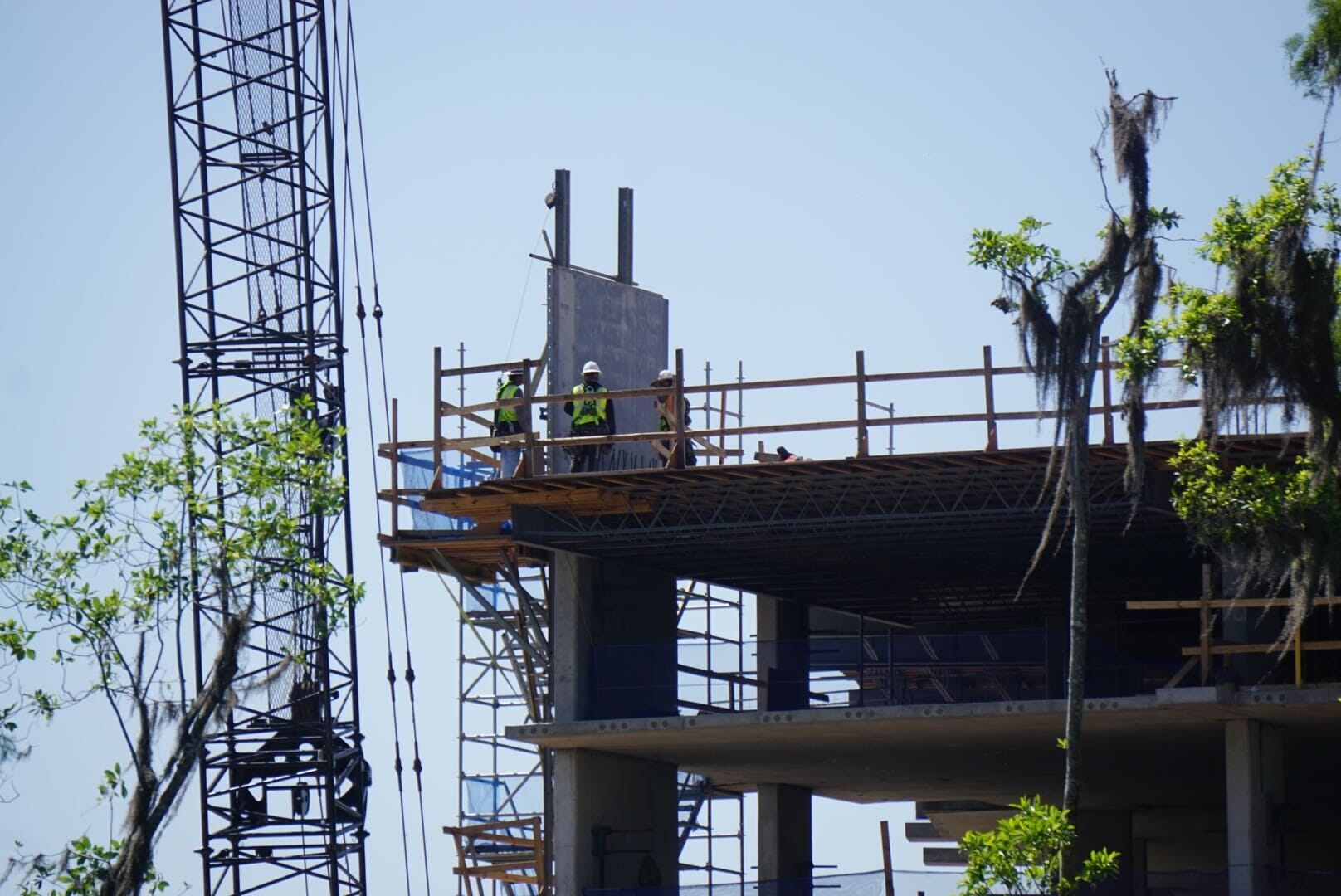 Construction workers at the Disney Lakeshore Lodge site are busy on the upper structure, with scaffolding and a crane in action. Surrounded by trees, this construction update highlights their progress amidst a scenic backdrop.