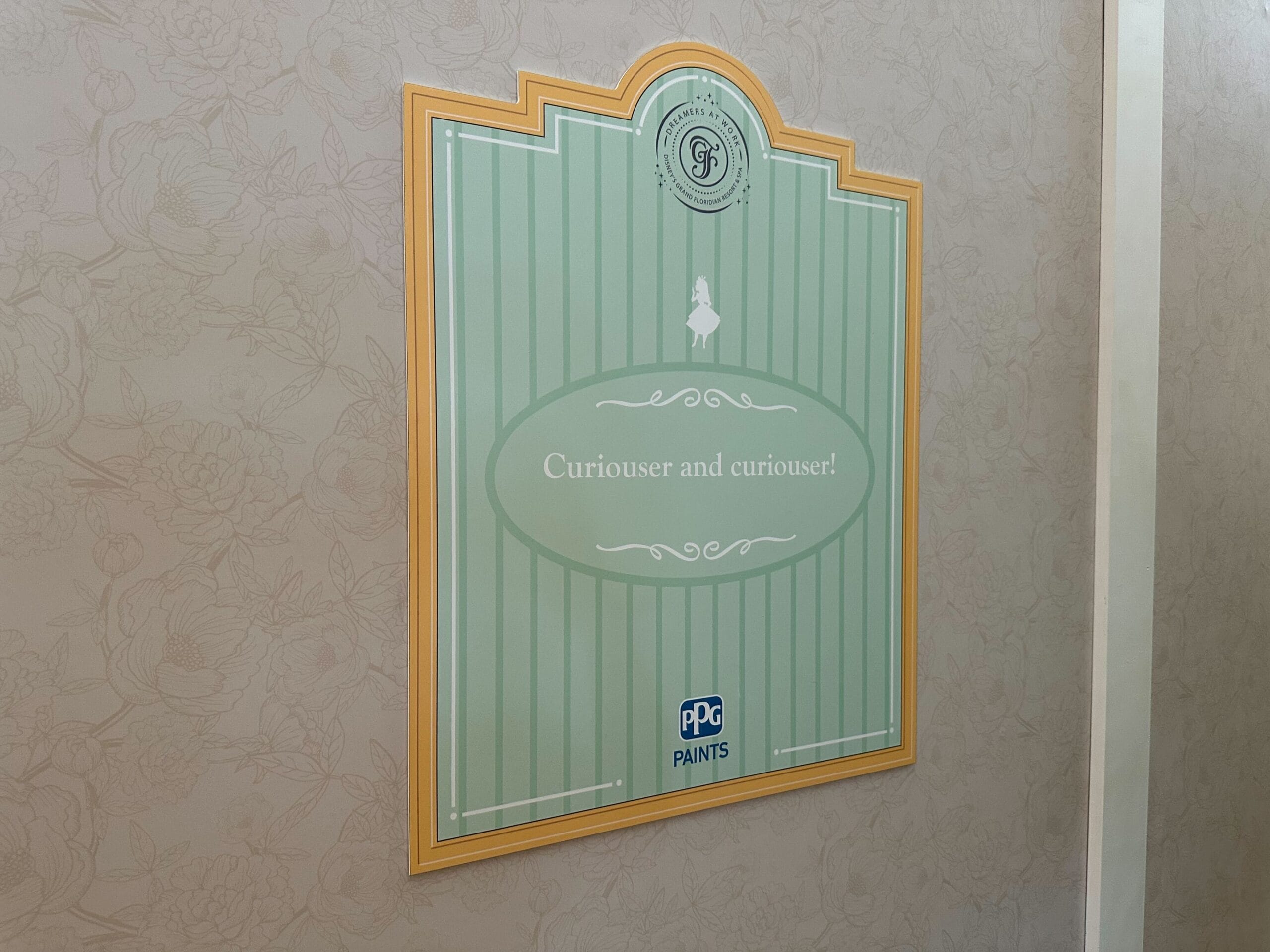 A green sign with white stripes reads Curiouser and curiouser!—part of new signage at Grand Floridian’s floral lobby wall.