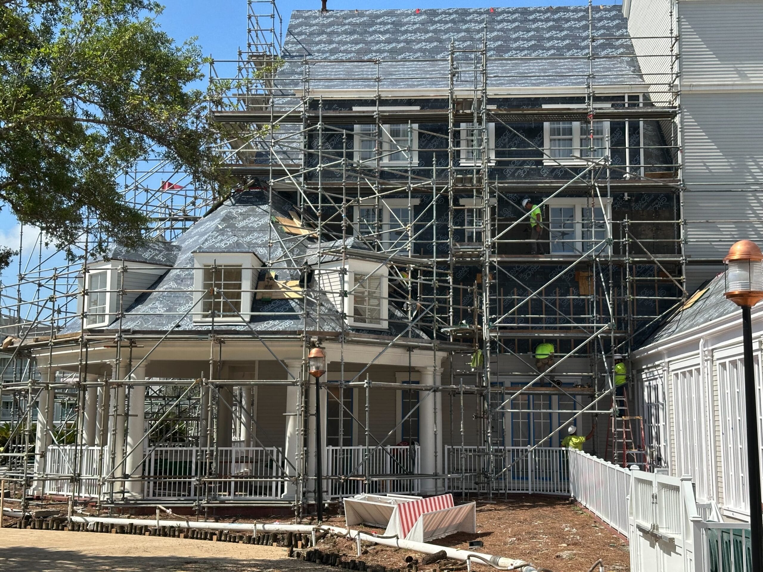 Scaffolding, reminiscent of a Tower of New Scaffolding, surrounds the multi-story white house undergoing renovations. To the left stands a tree, while construction materials are scattered in the foreground. It's as if Disney’s Yacht Club lent its elegance to this transformative scene.