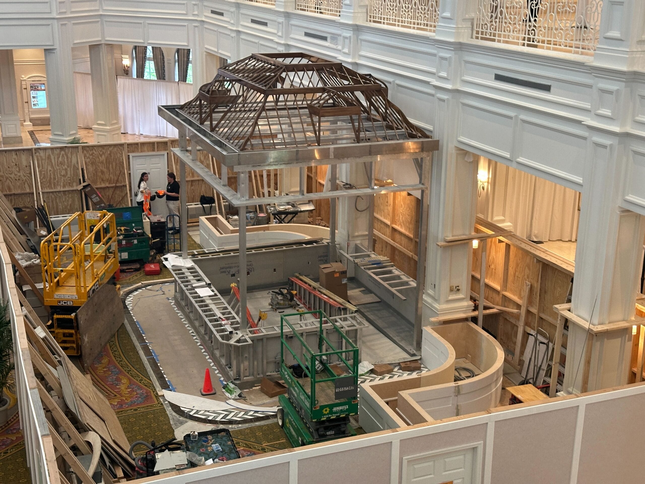 Indoor construction site with workers, scaffolding, and a partially built gazebo-like structure surrounded by white walls.