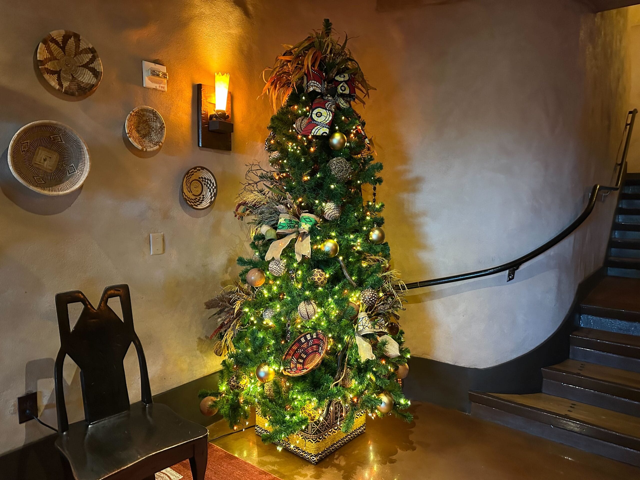 Decorated Christmas tree with lights stands in a warmly lit room beside a chair and a curved staircase, with decorative wall hangings.