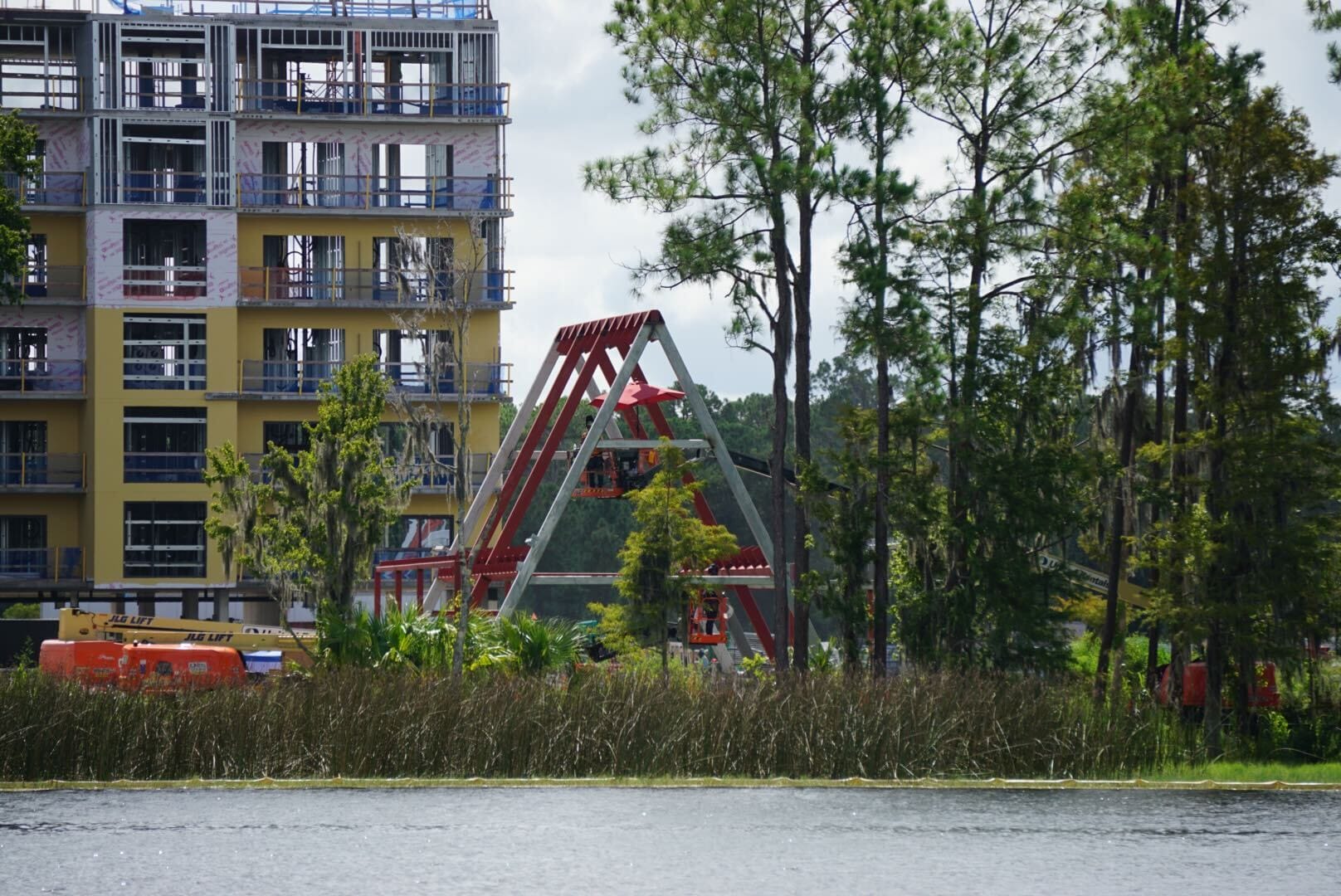 A construction site with a partially built building and a red swing ride near trees by a body of water.