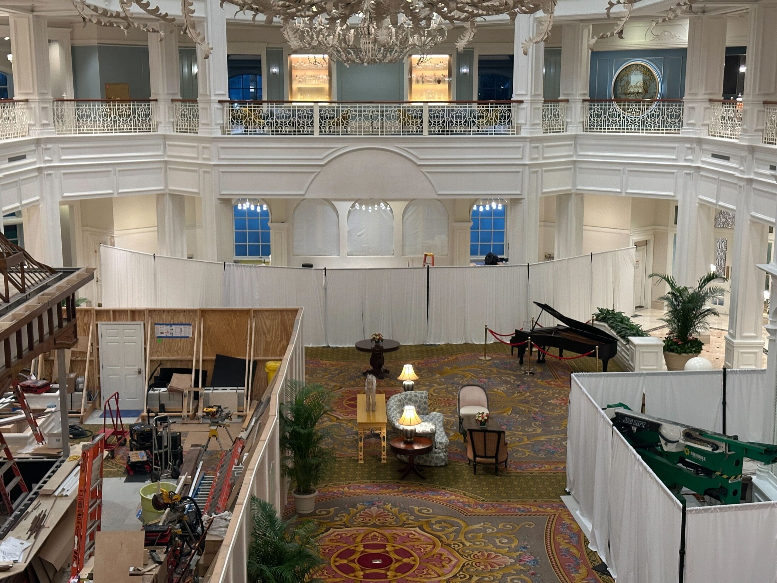 A grand hotel lobby with white walls, construction zones, covered furniture, and a piano separated by white partitions.