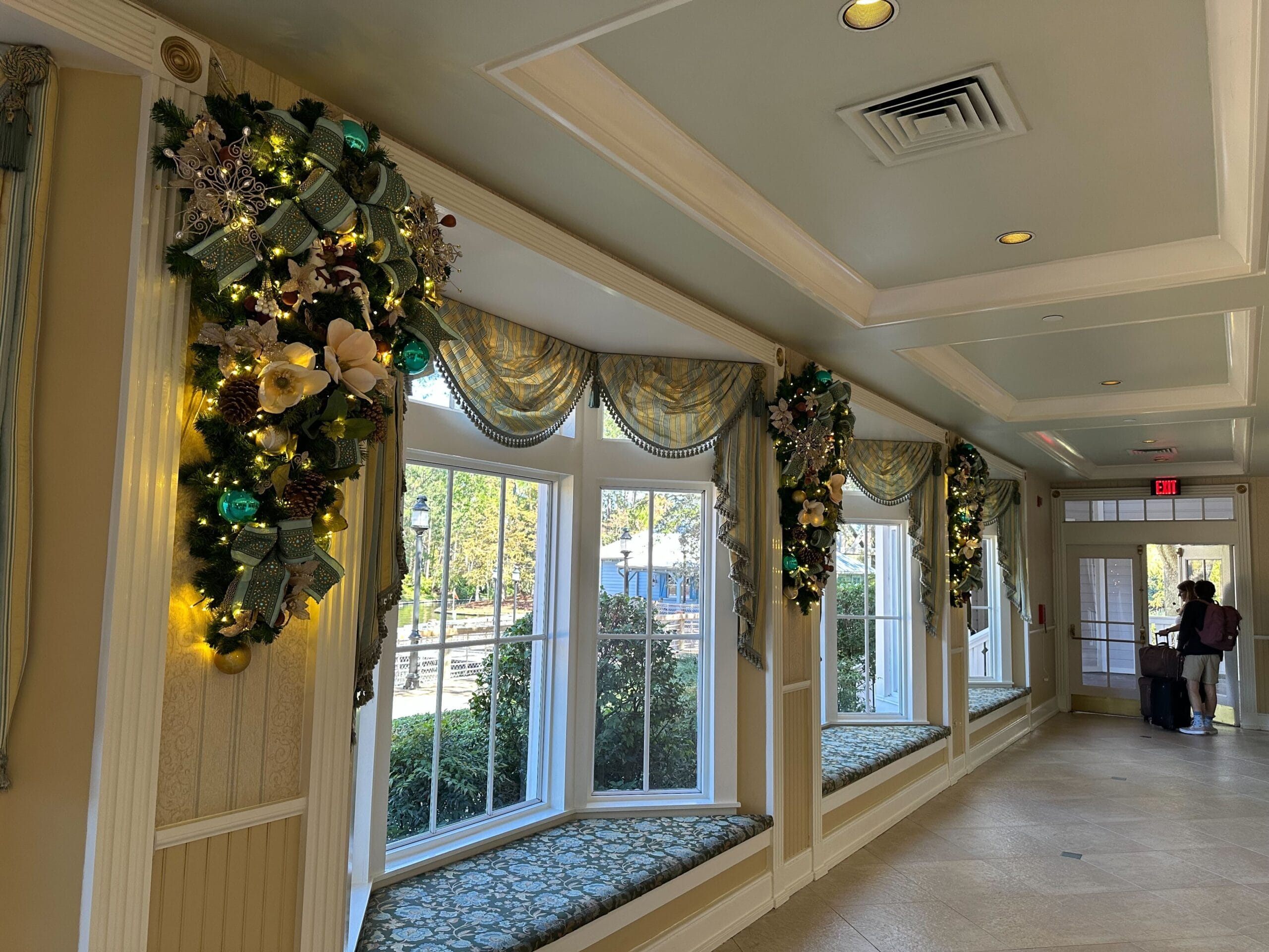 A hallway with large windows is decorated with festive garlands and ornaments. Natural light illuminates the beige walls and patterned cushion seating.
