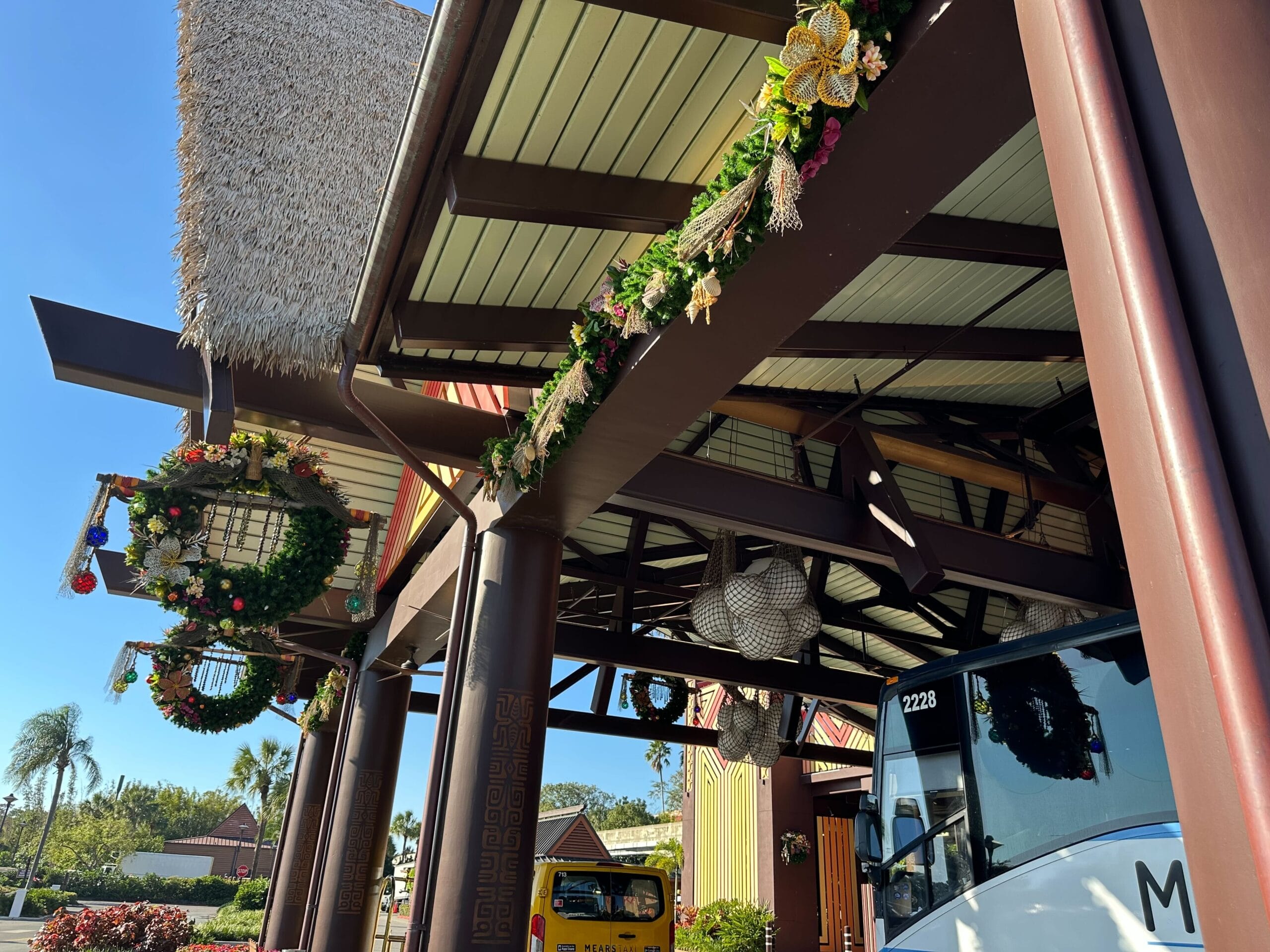 Decorated entrance with floral garlands and wreaths, featuring tall columns and a thatched roof. A bus is partially visible in the foreground.