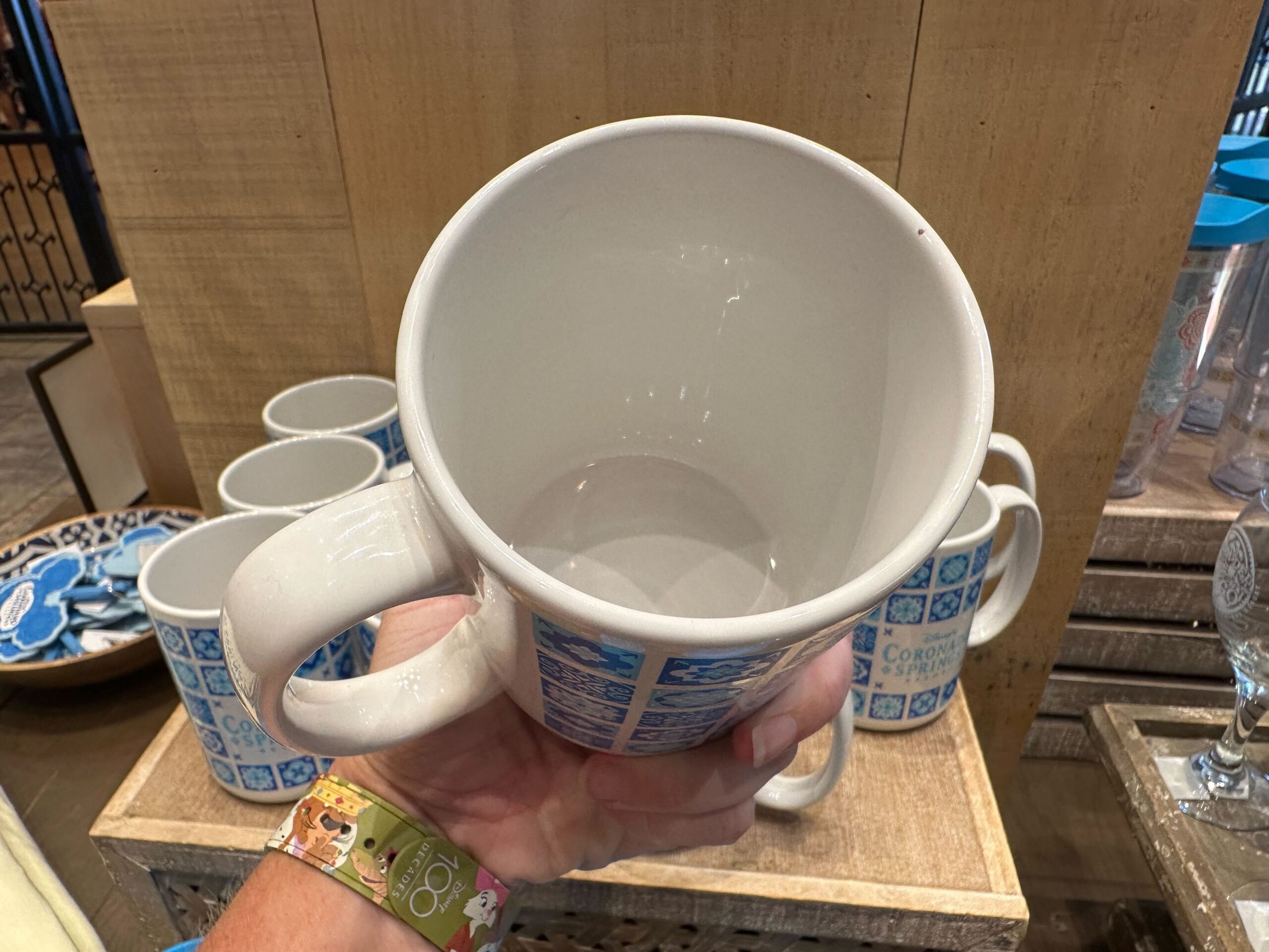 A hand holds a large white mug with a blue pattern among several similar mugs on a wooden display shelf, showcasing delightful merchandise at Coronado Springs.