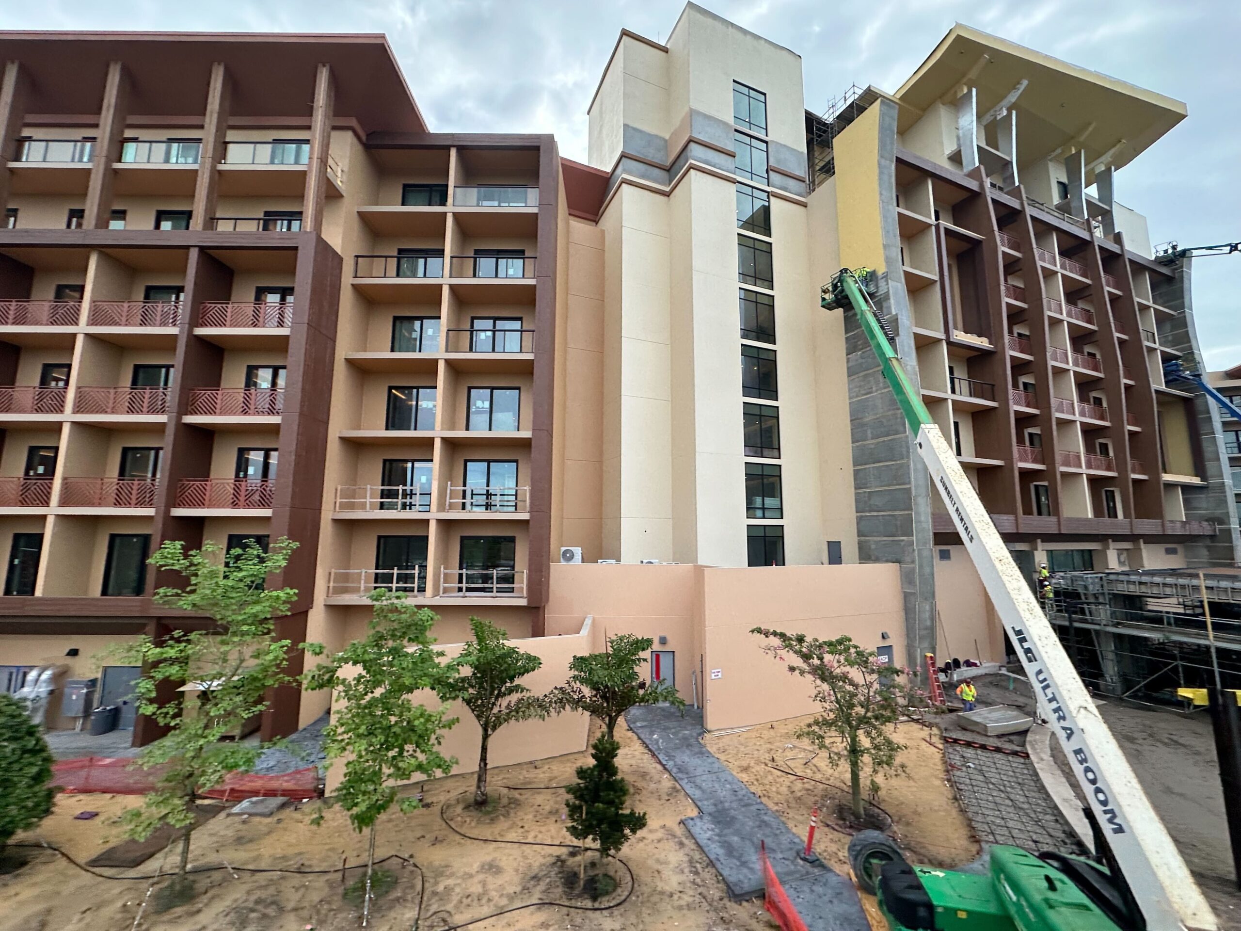 A multi-story building under construction with a crane elevating materials. The structure has various balconies and windows, and there is scaffolding on the right. Small trees and a path are in the foreground.