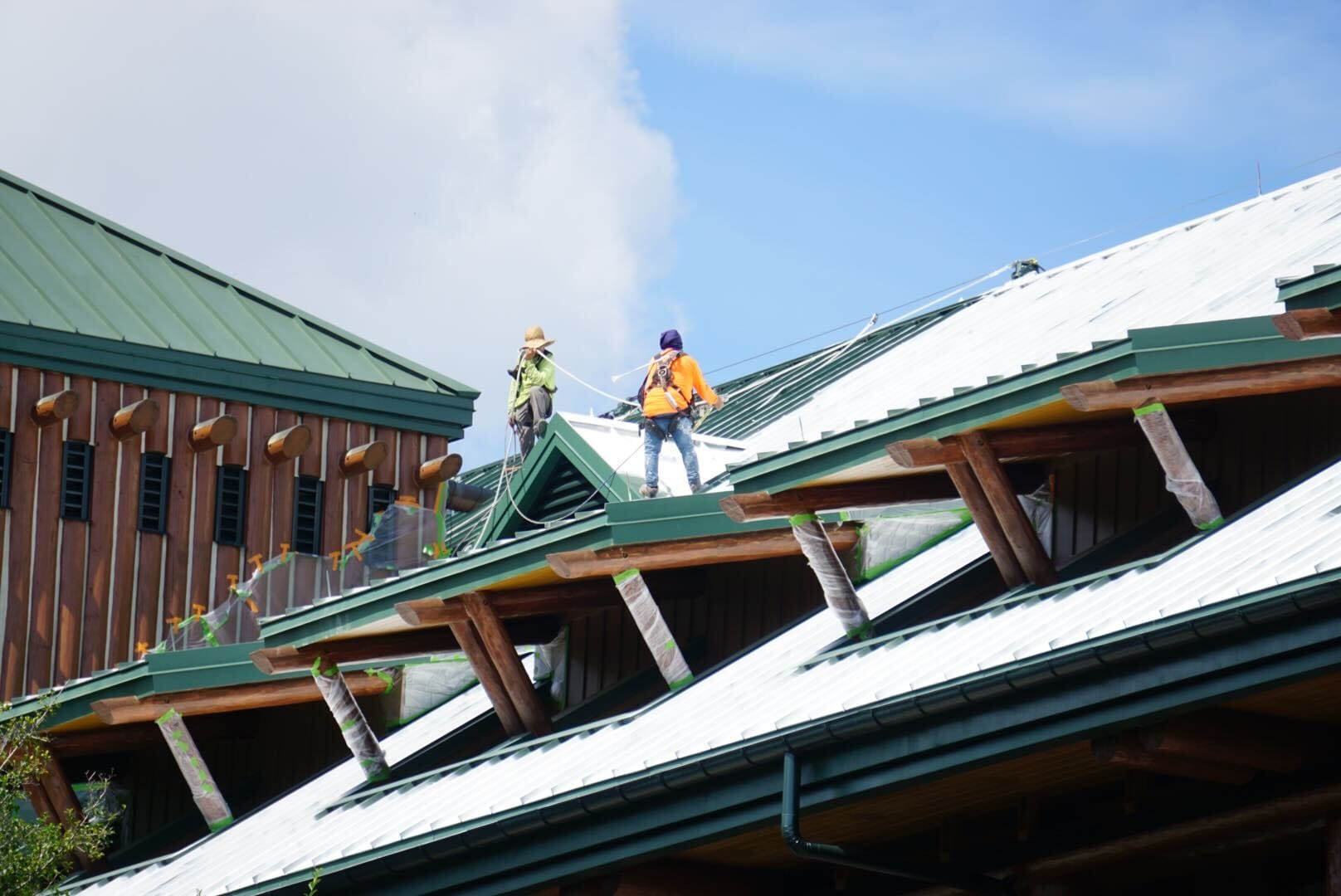 Two workers in safety gear stand on a green metal roof under a blue sky, near a wooden building.