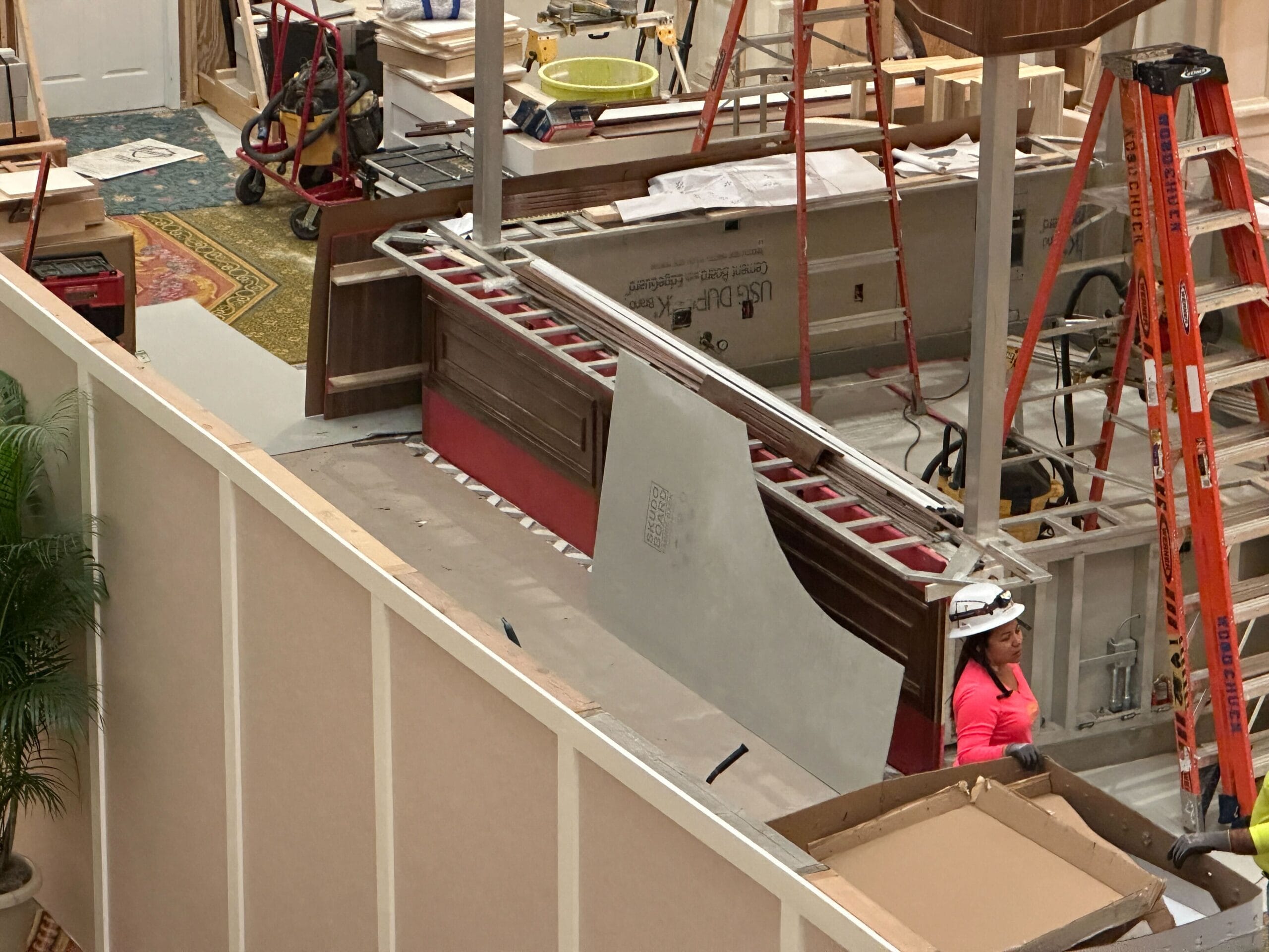 Woman in a pink shirt and hard hat works on an indoor Grand Floridian construction site with ladders and building materials.