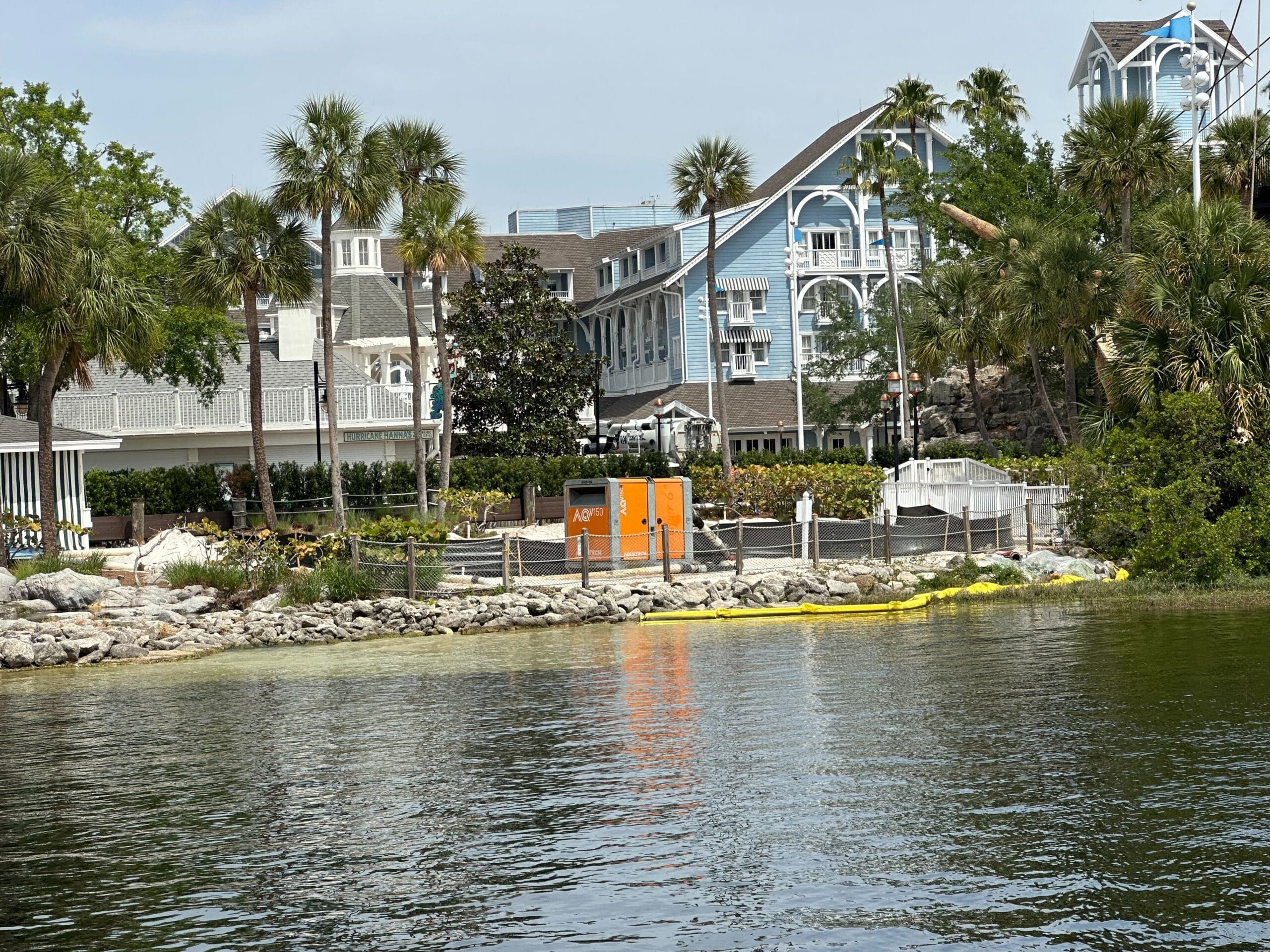 The waterfront area, featuring a blue-roofed building reminiscent of Disney's Yacht Club, is adorned with palm trees and dotted with scaffolding and construction barriers near the shoreline.