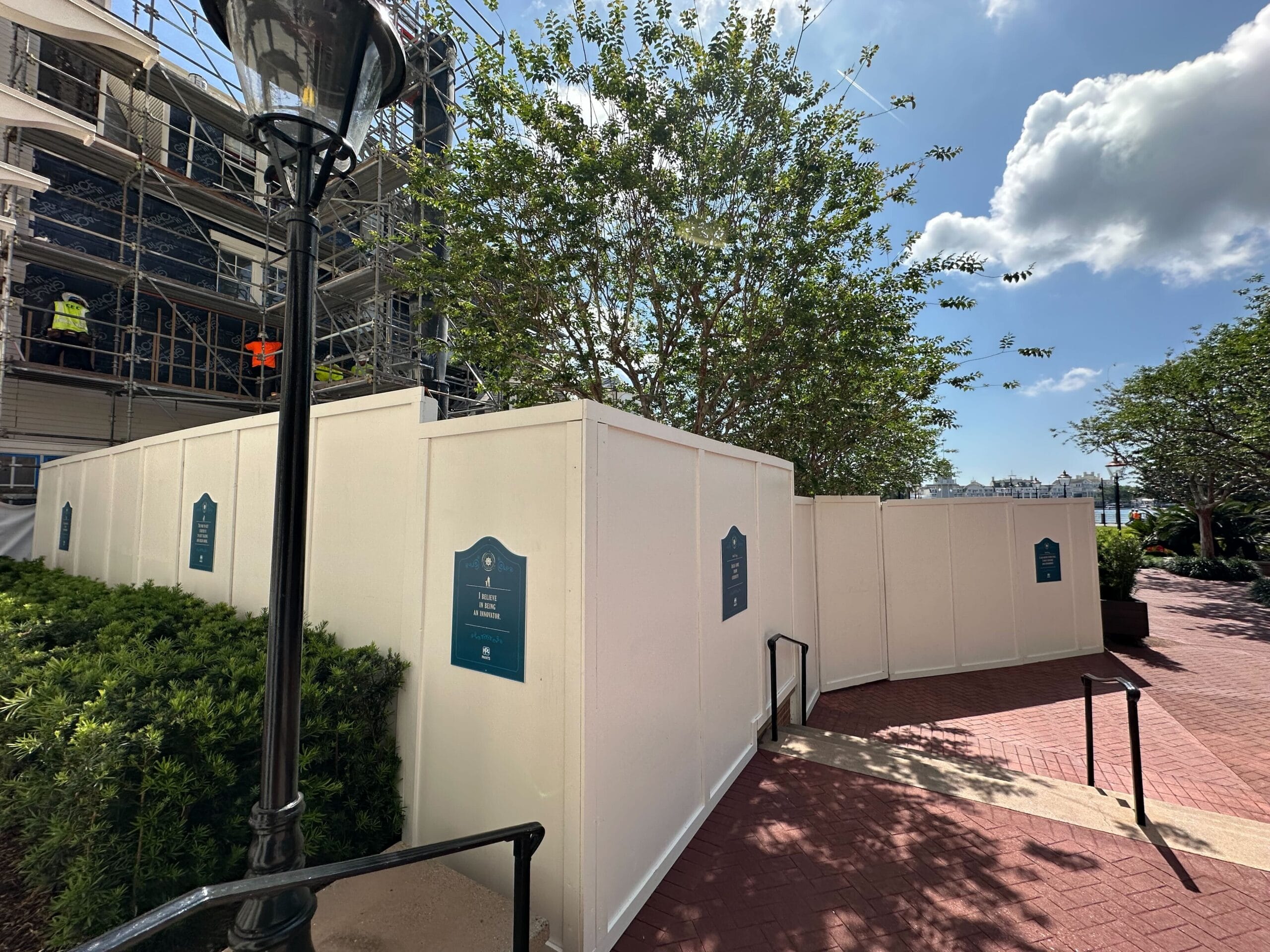 A tower looms behind the construction site, where scaffolding peeks over white barriers. Surrounded by trees and shrubs, the scene unfolds under a partly cloudy sky, with a lamp post standing in the foreground, reminiscent of Disney’s Yacht Club ambiance.