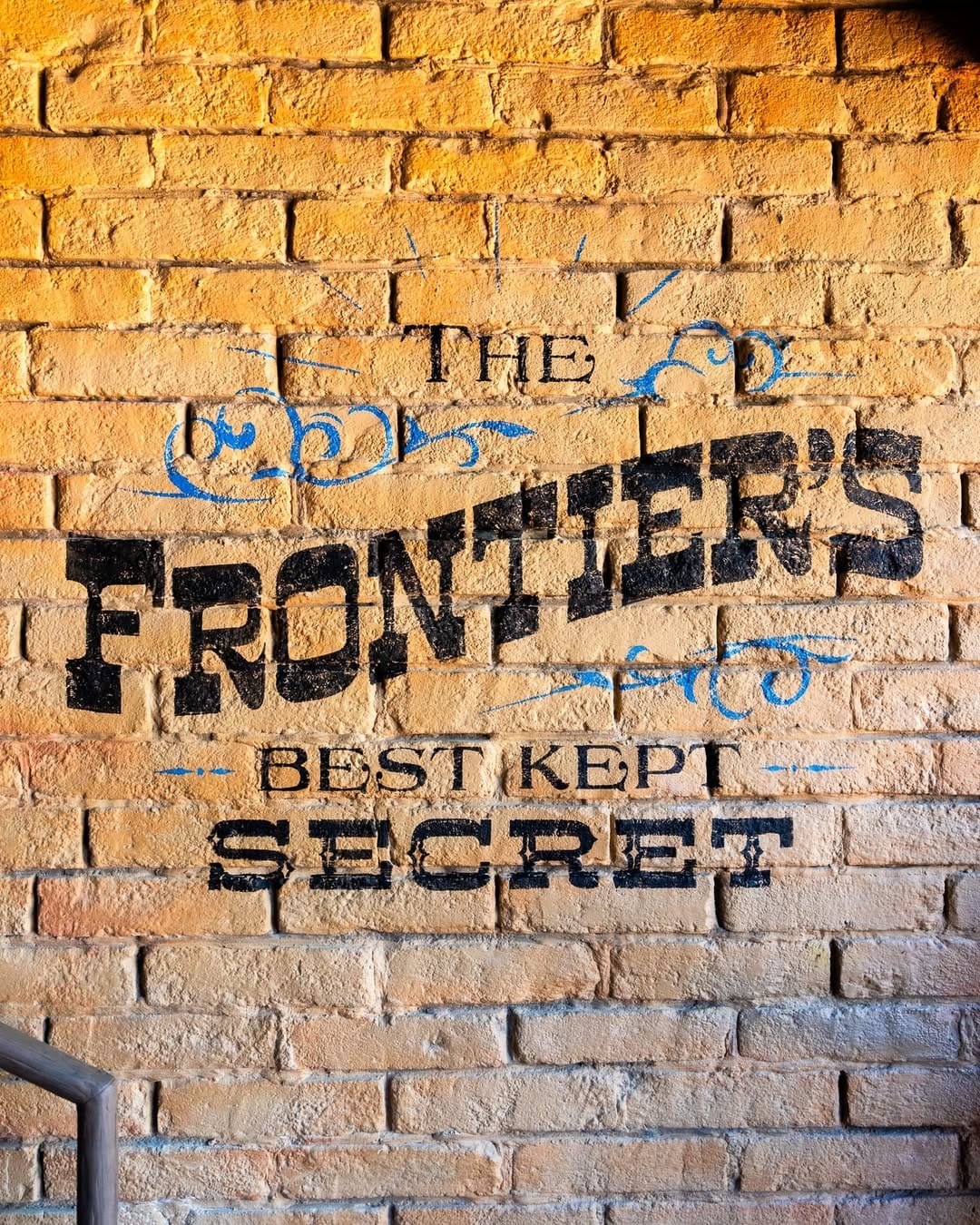 Text on a brick wall reads The Frontier's Best Kept Secret in decorative black and blue fonts.