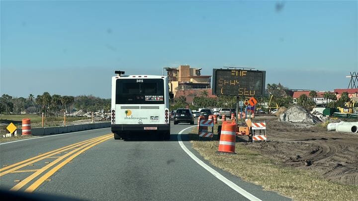 Traffic on a two-lane road near Walt Disney World is lined with orange construction barrels and a digital road sign displaying estimated wait times, as cars queue behind a bus.