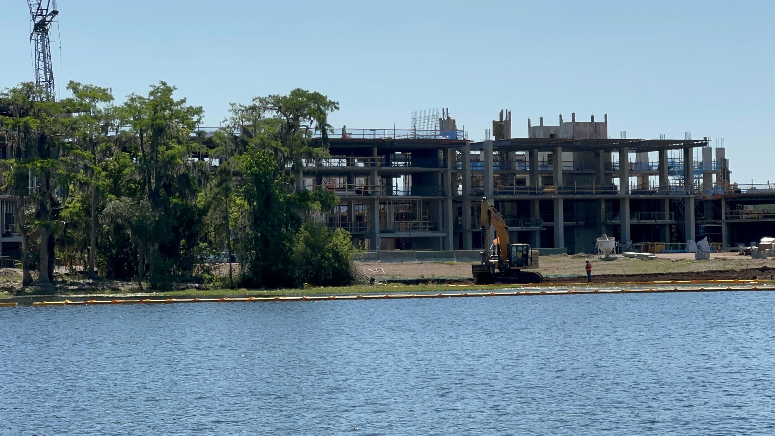 A multistory Disney Lakeshore Lodge is under construction near a body of water, with a crane and machinery visible. Trees partially obscure the view on the left side.