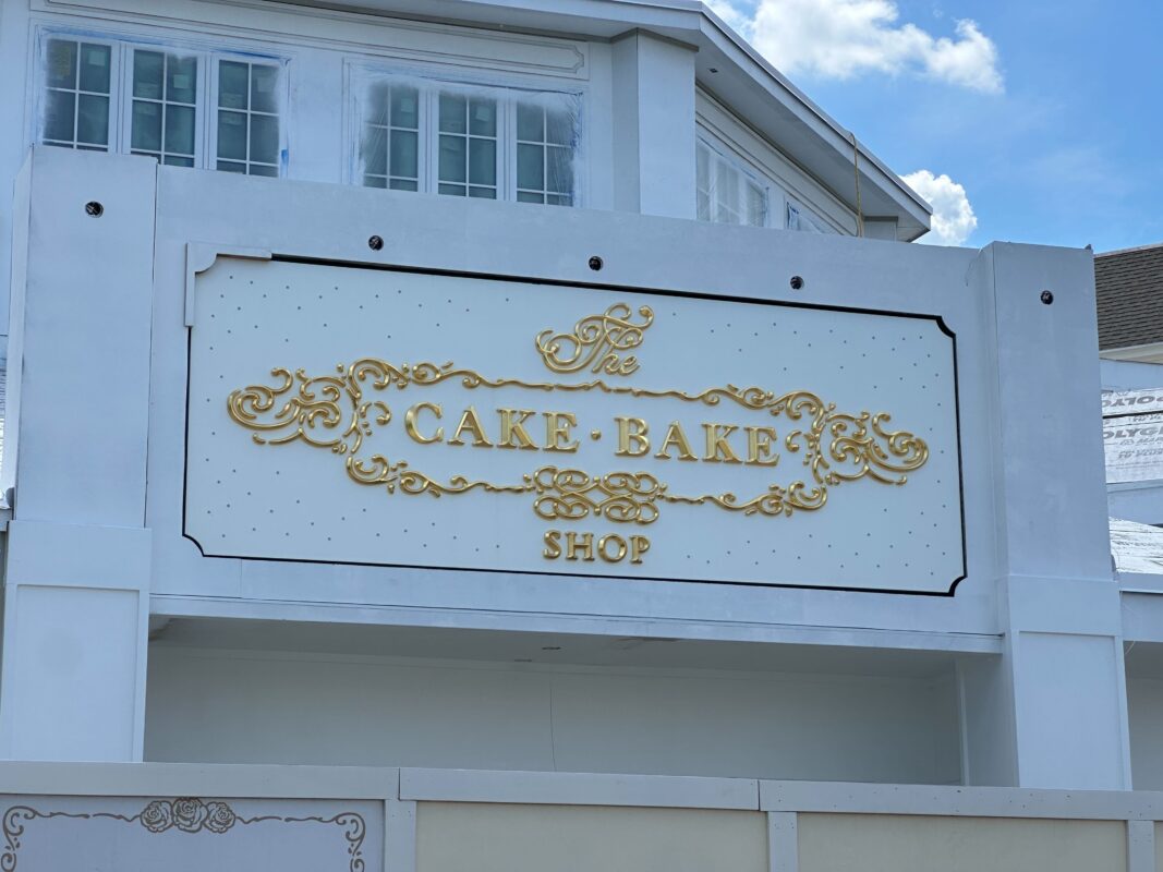 A white building with a sign that reads The Cake Bake Shop in ornate gold lettering stands next to an Auto Draft station.