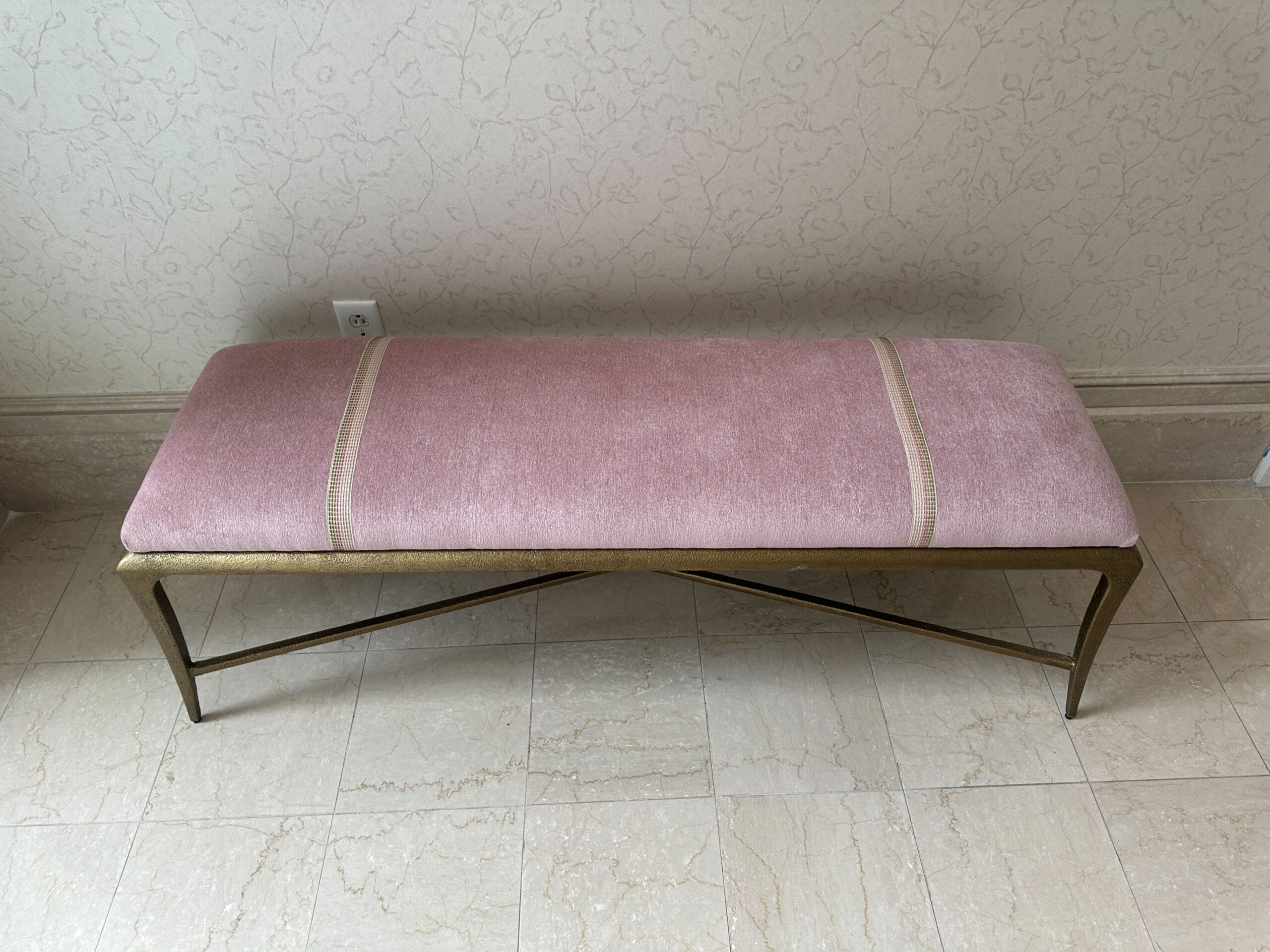 A pink upholstered bench with gold legs sits on a tiled floor against a patterned cream wall.