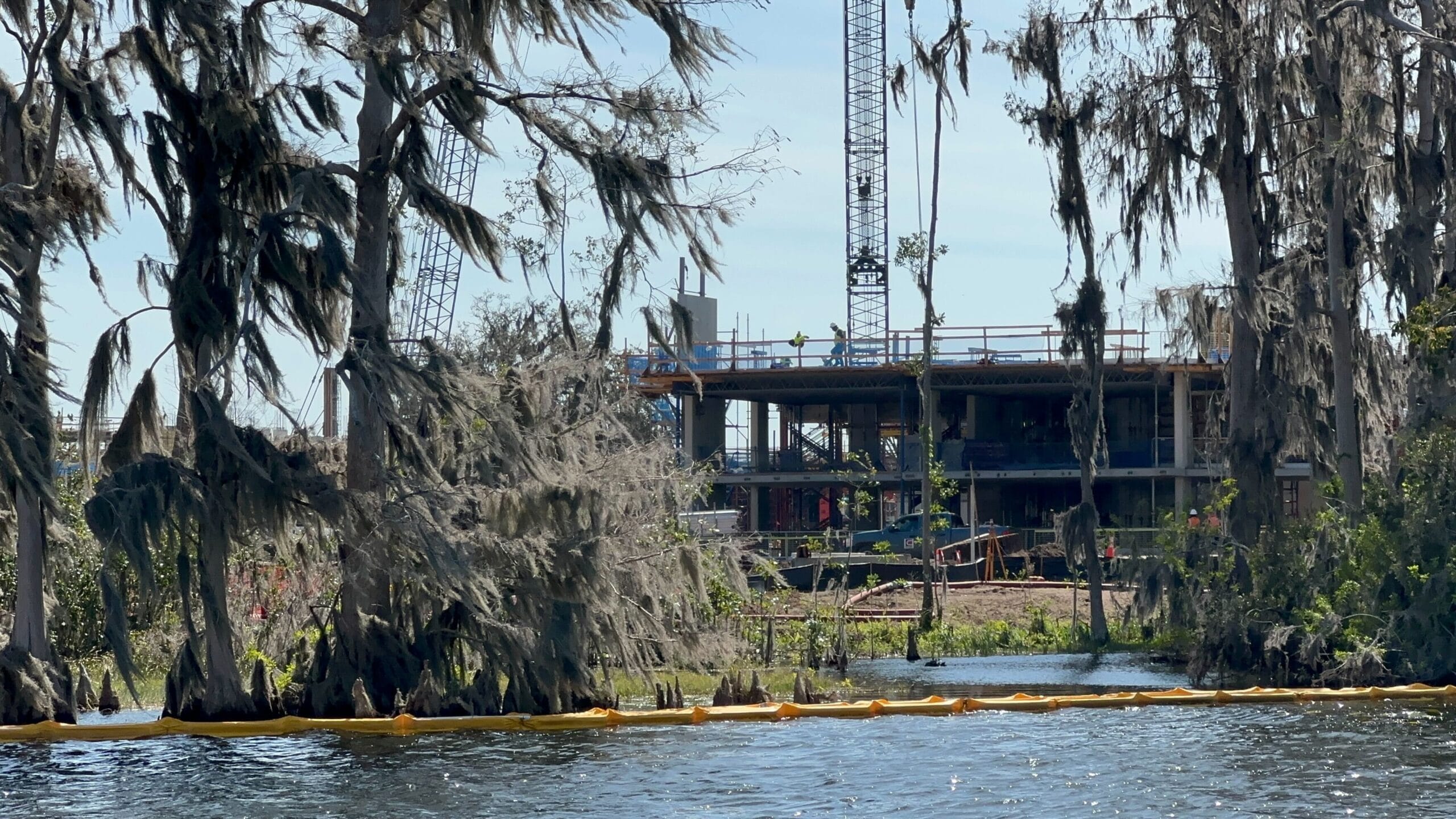Construction site by a lake with cranes and partially built structures, surrounded by trees draped in moss. A yellow barrier floats on the water, as the nearby beach is blocked for construction of the future Lakeshore Lodge.
