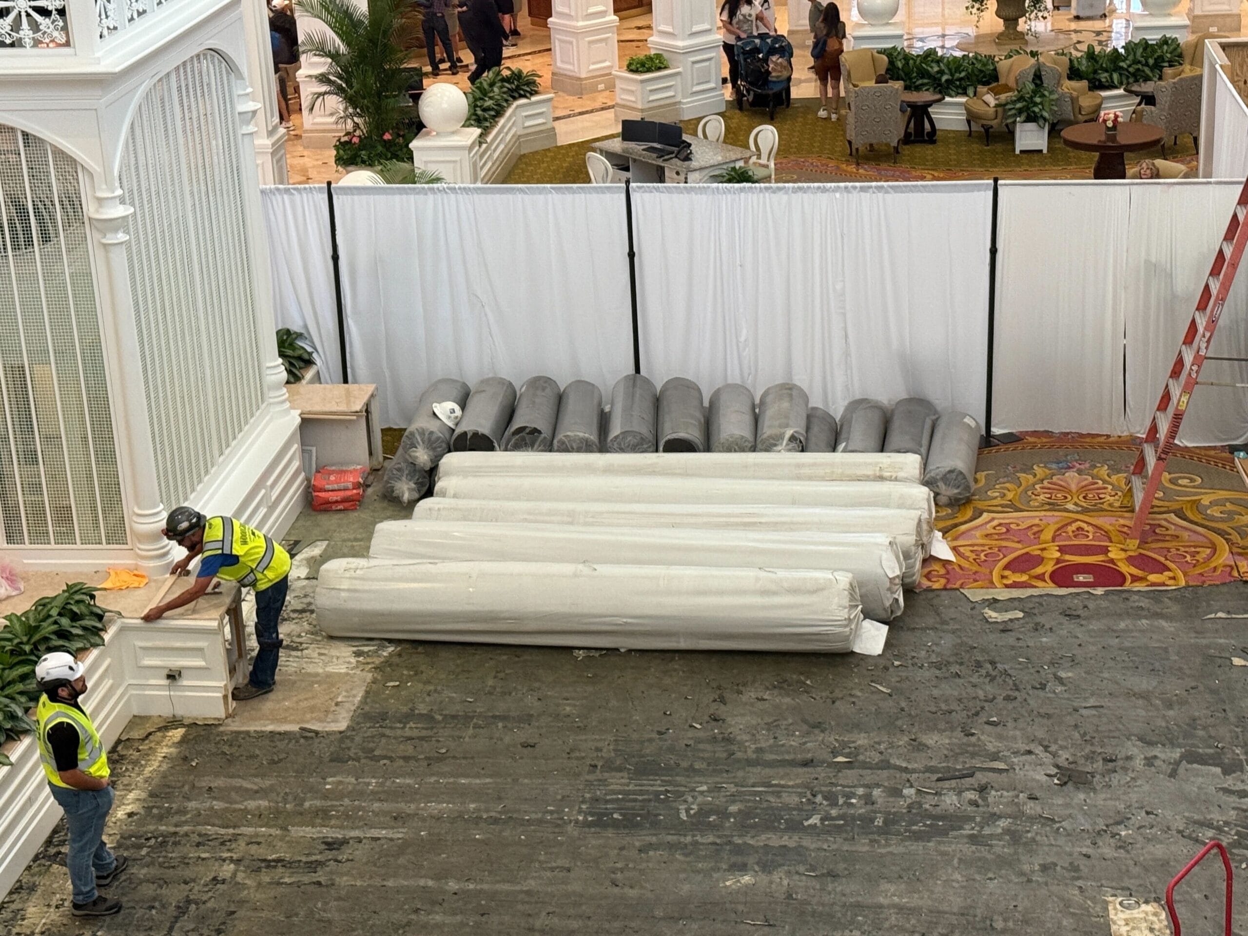 Two construction workers stand near rolled-up carpets and columns in a partially renovated indoor area with white barriers.