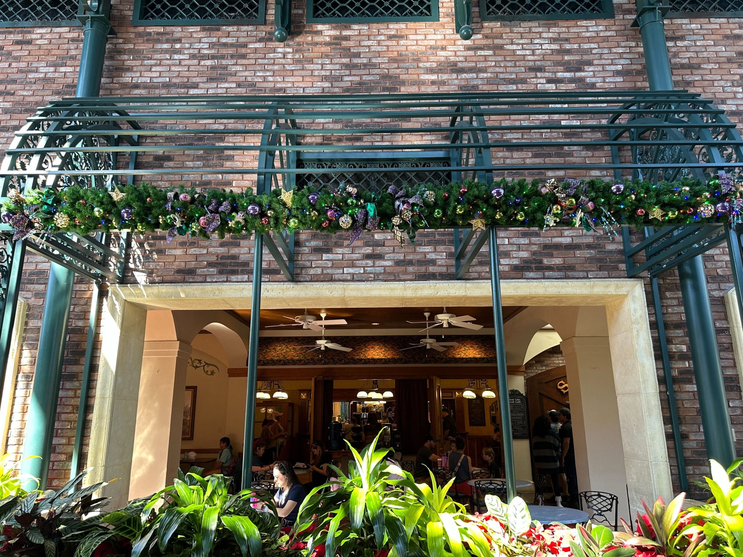 Brick facade with green pergola and floral garland above an open-air cafe. Indoor seating visible through large arches. Bright plants in the foreground.