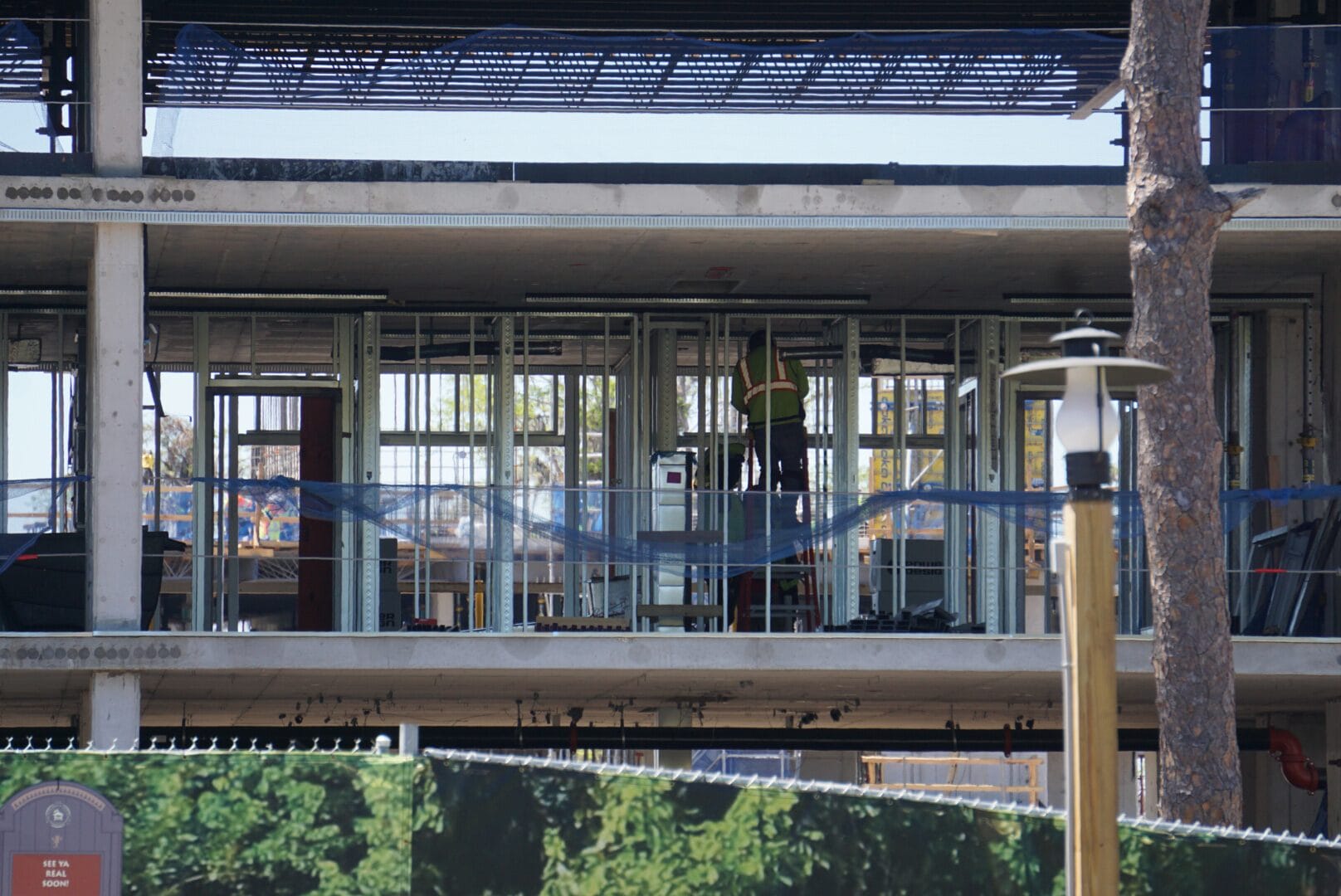A person in safety gear works on the framework of a multi-story building under construction, surrounded by metal beams and scaffolding.