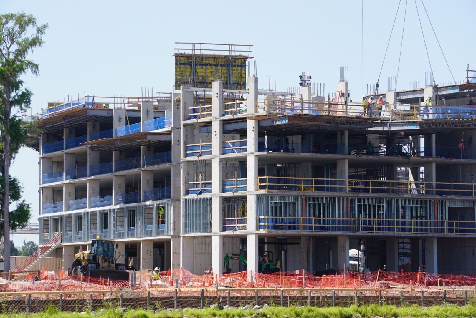 A multi-story building under construction rises near the lakeshore, with scaffolding and construction materials scattered around. A bulldozer is parked nearby, and orange safety netting surrounds the site like a temporary lodge in progress.