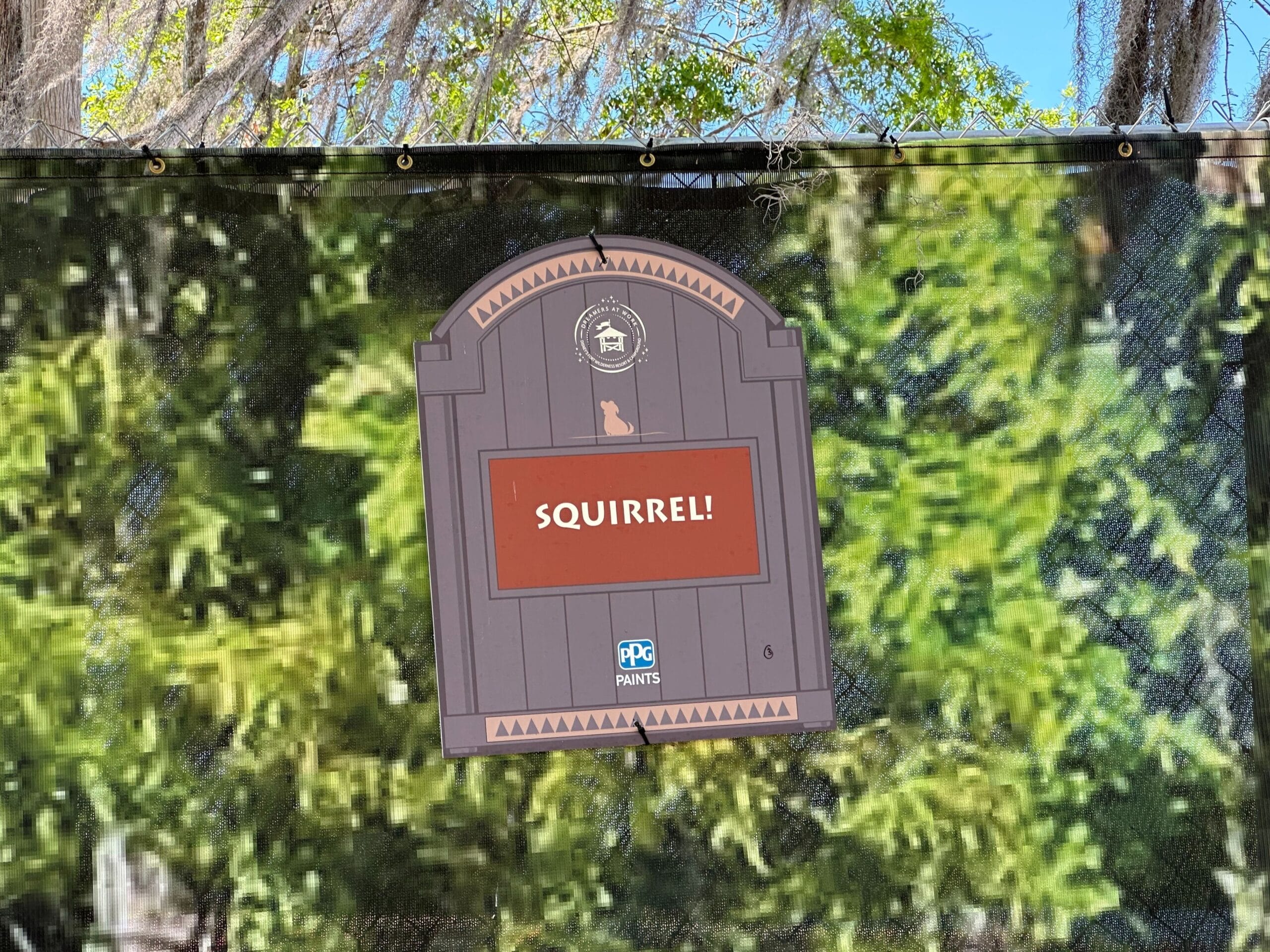 Sign with the word SQUIRREL! in white on red, attached to a wooden gate design, set against a backdrop of green foliage and a black mesh fence.