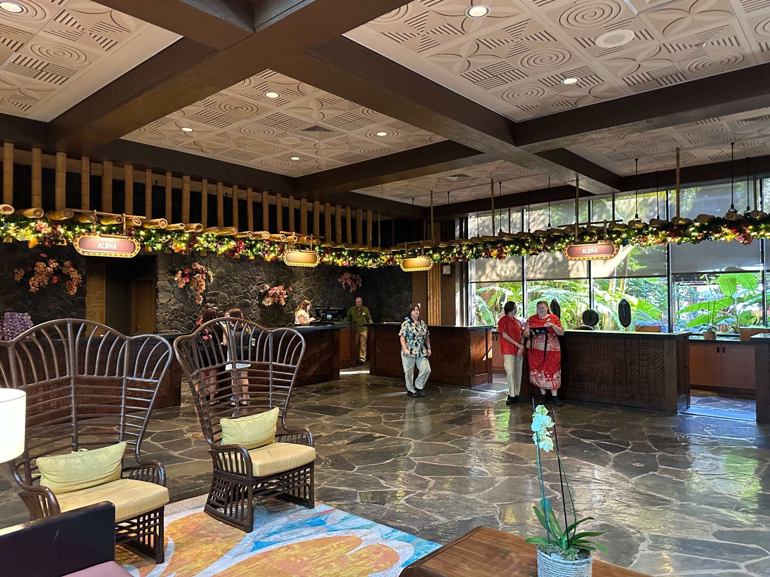Hotel lobby with floral decorations, rattan chairs, and people checking in at the front desk under soft lighting.
