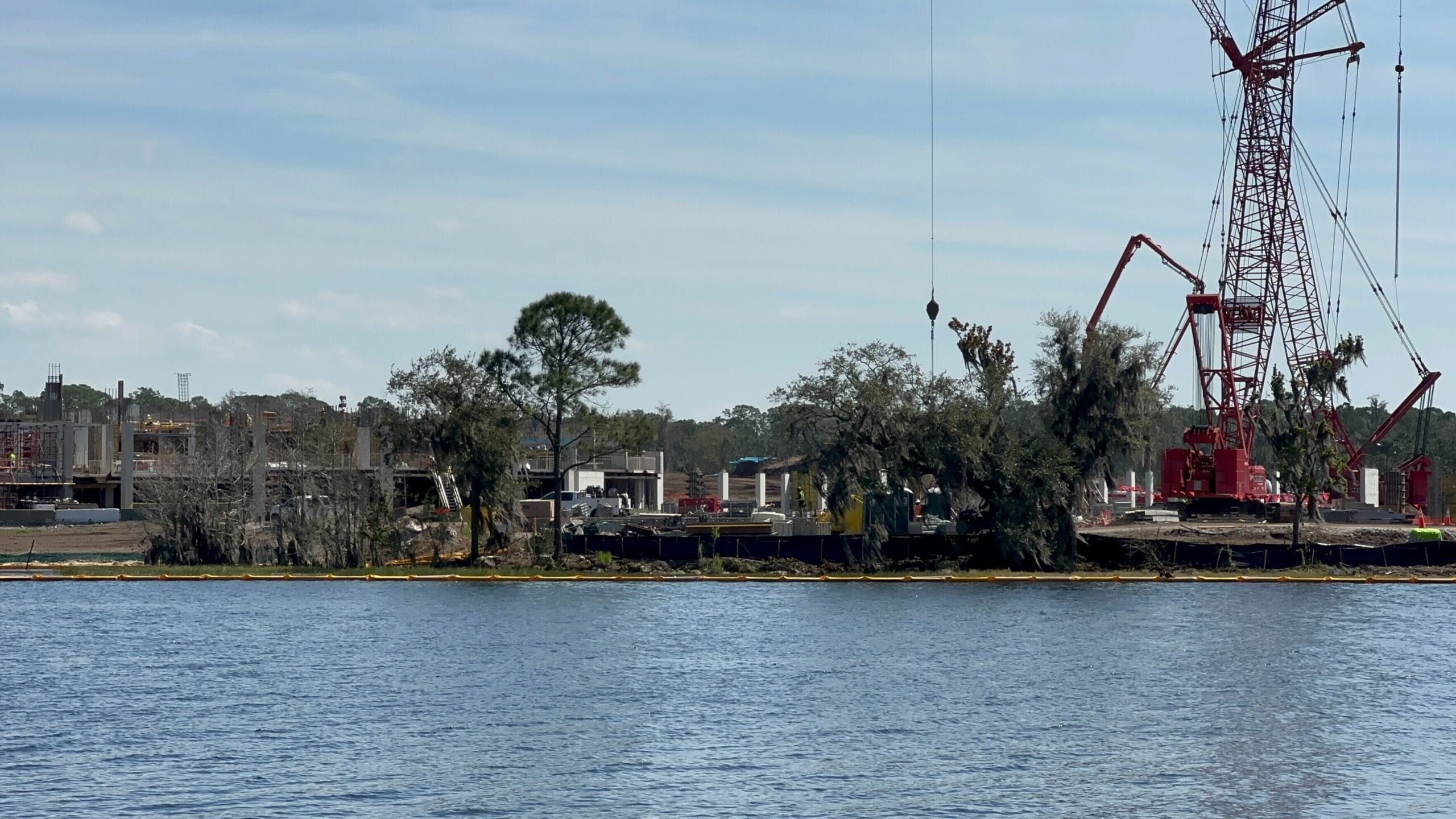 A large red crane operates near construction materials by a waterfront at Lakeshore Lodge Construction, with trees and buildings in the background under a clear sky.