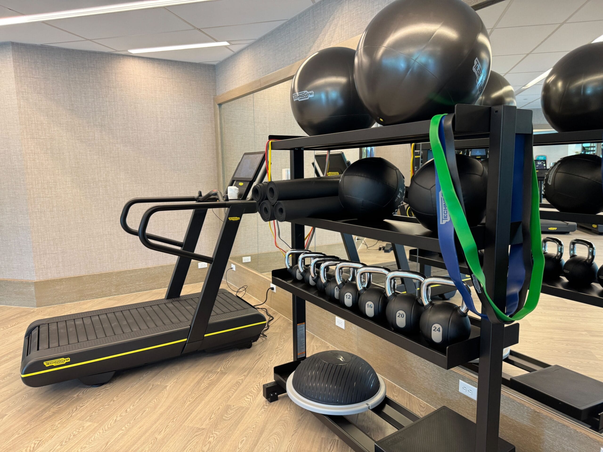 A gym interior with a treadmill, exercise balls, kettlebells, resistance bands, and a balance trainer on a wooden floor.