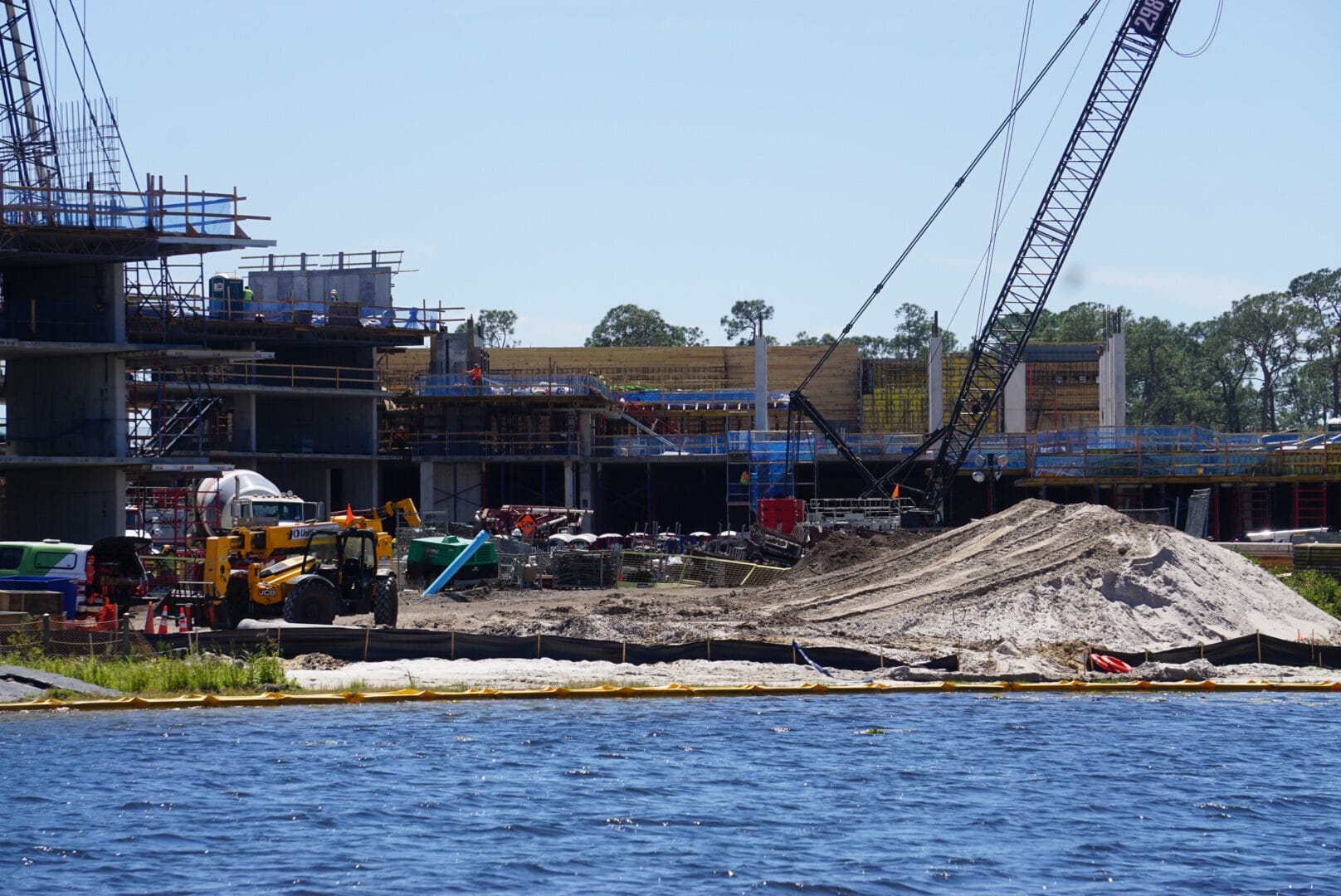 Construction site by a body of water, featuring cranes, building materials, and sand piles. Multiple structures are partially built under a clear sky.