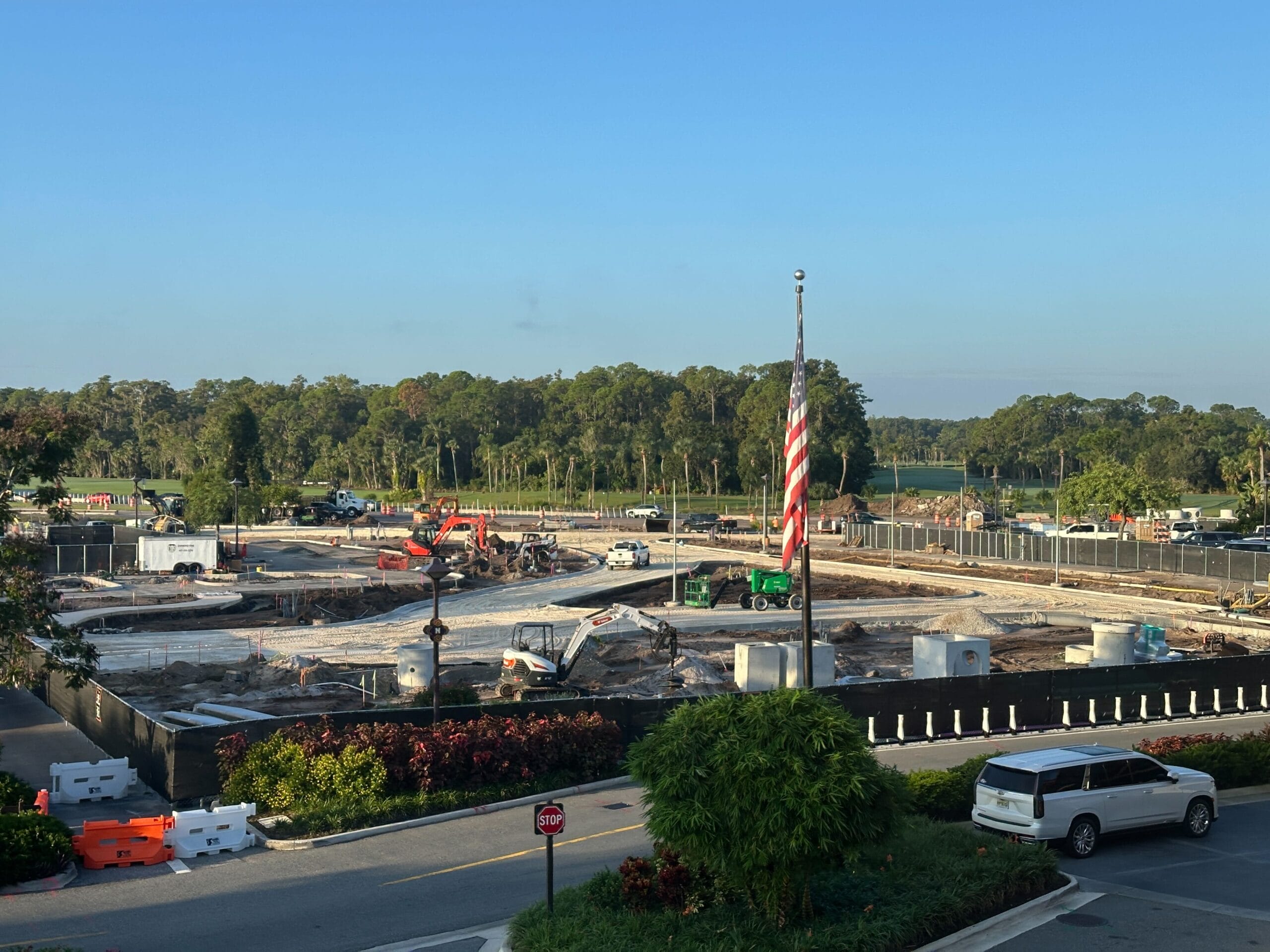 Construction site with vehicles and workers, American flag in foreground, trees and cars in the background under a clear sky.