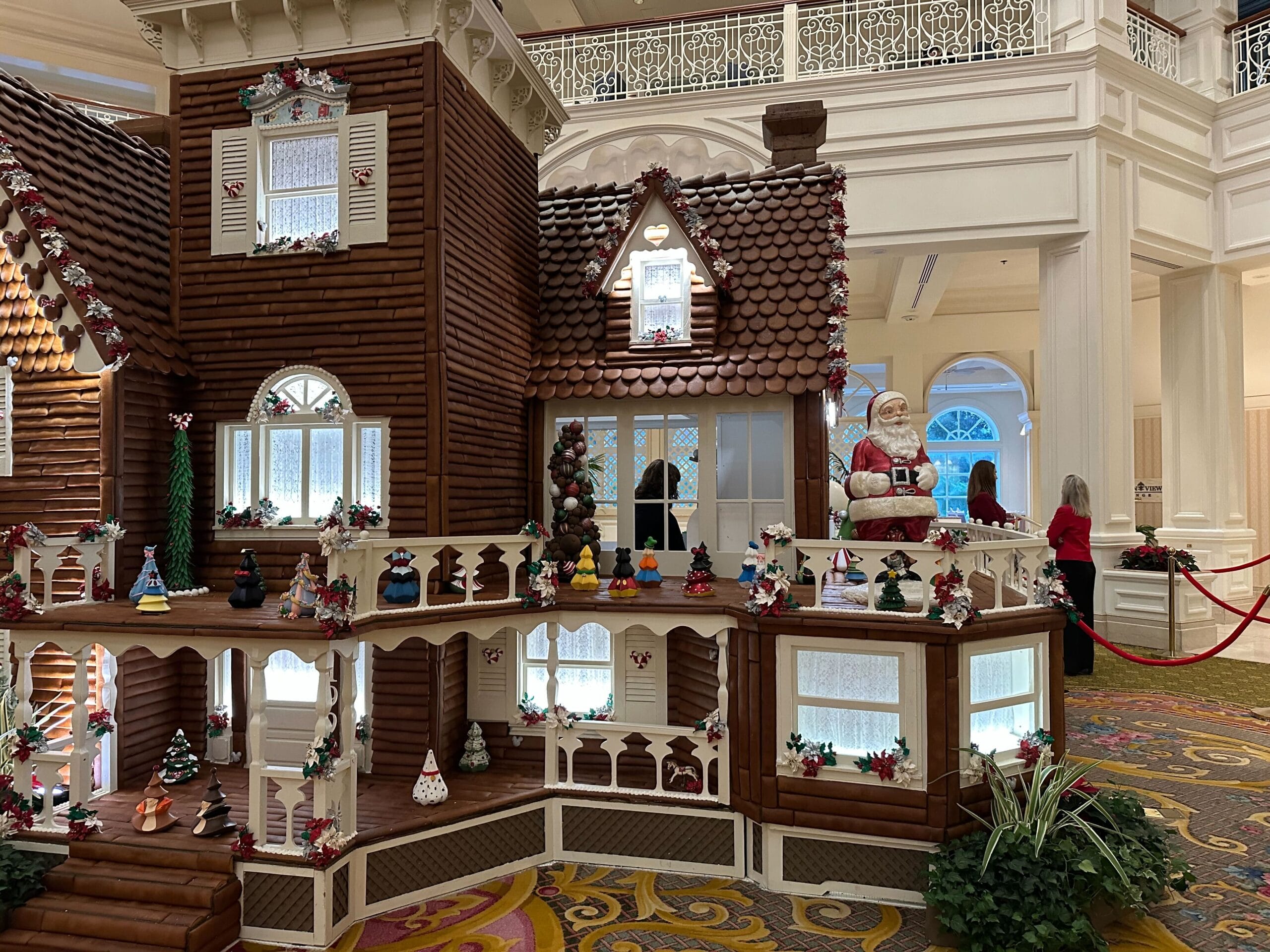 Large gingerbread house display with Santa Claus figure, festive decorations, and small ornaments in an ornate indoor setting.