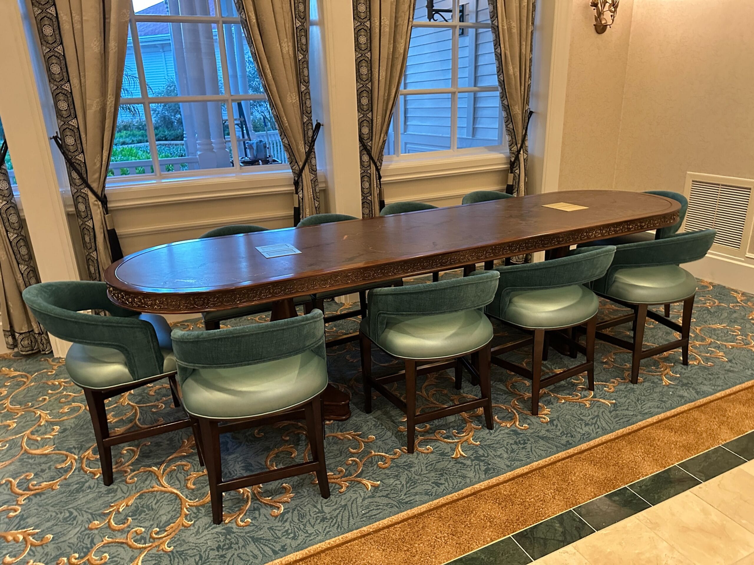 A long wooden table with eight teal chairs in an elegant Grand Floridian room with large windows and patterned carpet.