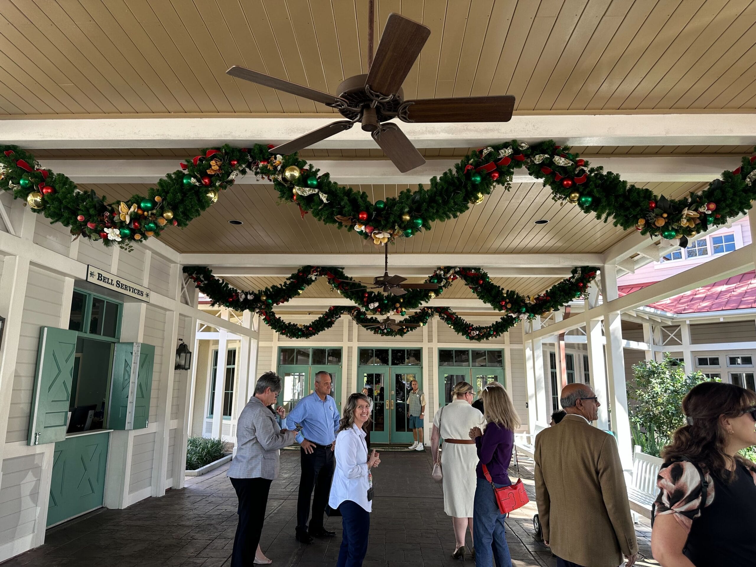 A group of people stand under a decorated entrance with green garlands and gold and red ornaments. A ceiling fan is above them.