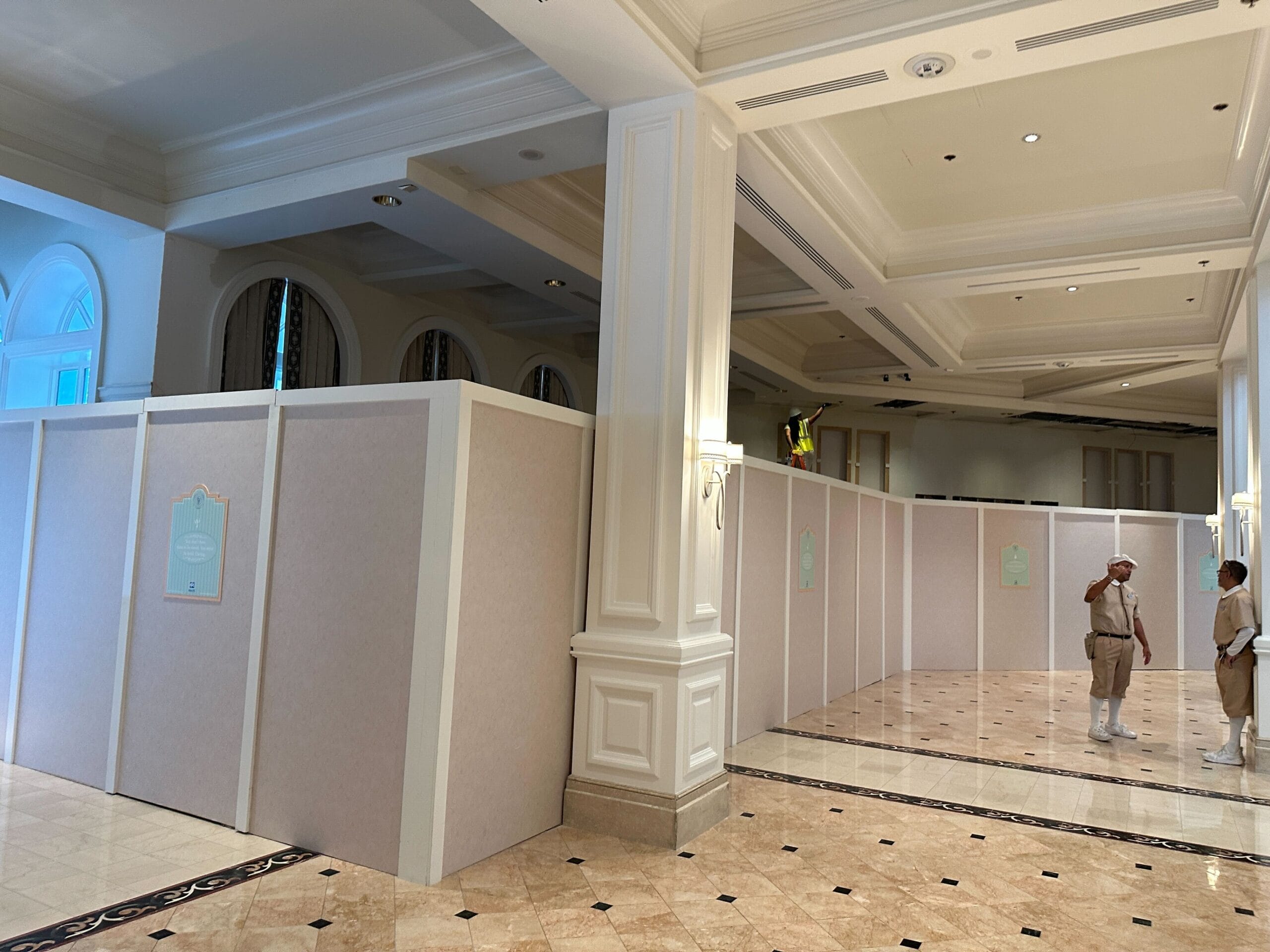 Two workers stand near tall construction walls inside an elegant, columned building with marble floors.