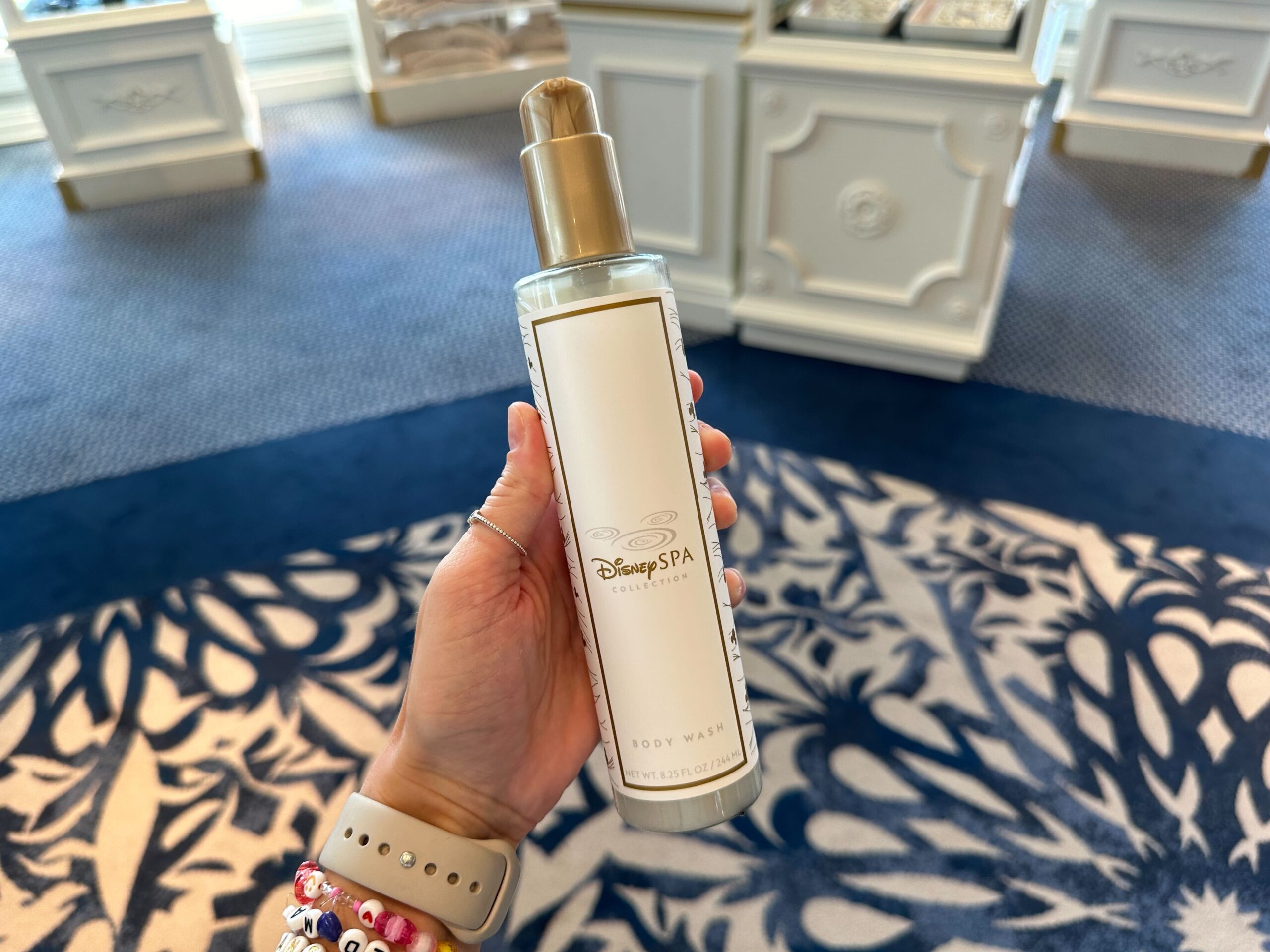 A hand holding a bottle of body wash labeled Disney Spa Collection against a carpeted background with furniture, reminiscent of the elegant Grand Floridian style, introduces exciting new merchandise to elevate your self-care routine.