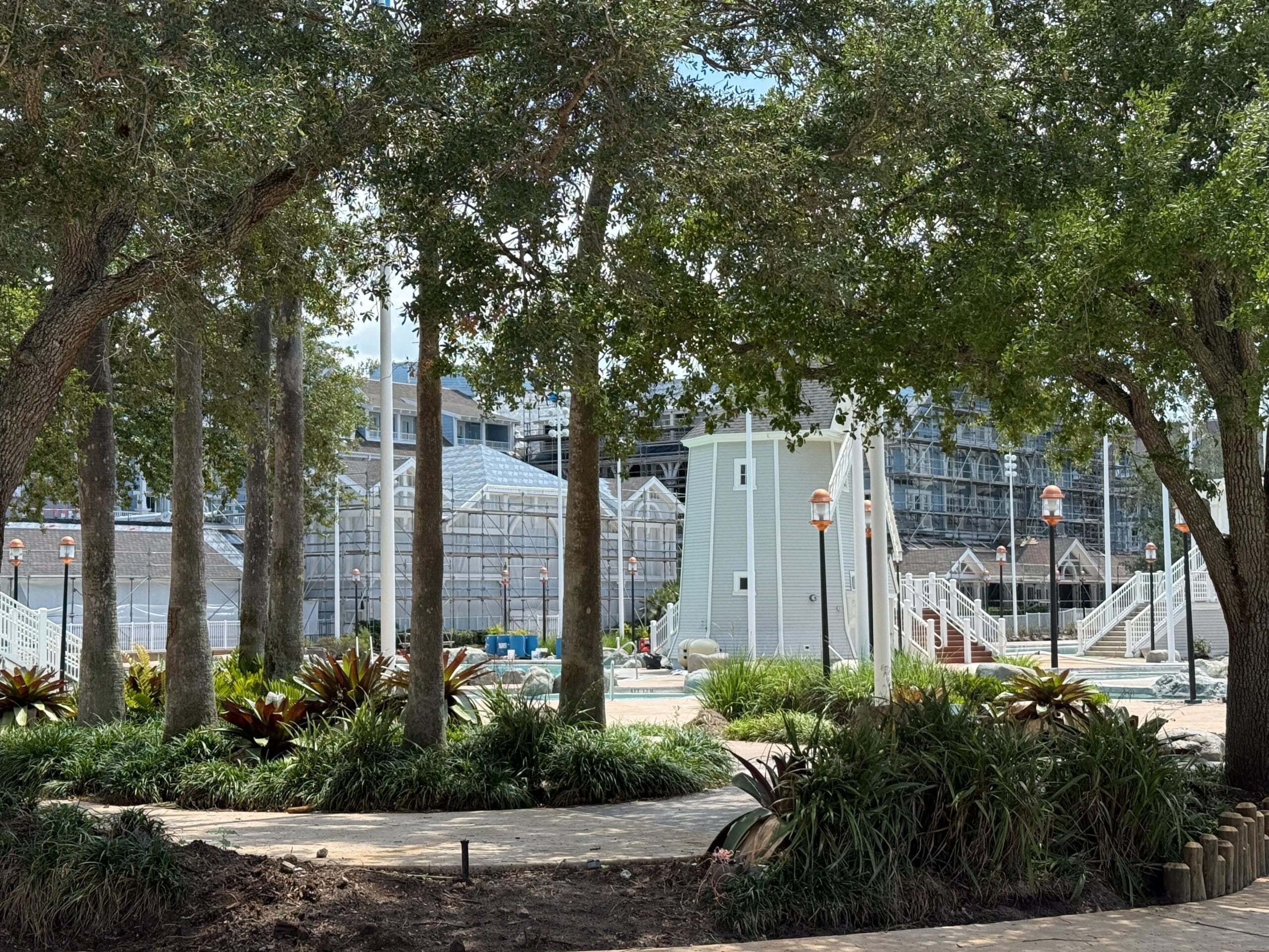 Greenhouses and a light gray building under construction, partially obscured by trees and landscaped plants in the foreground.