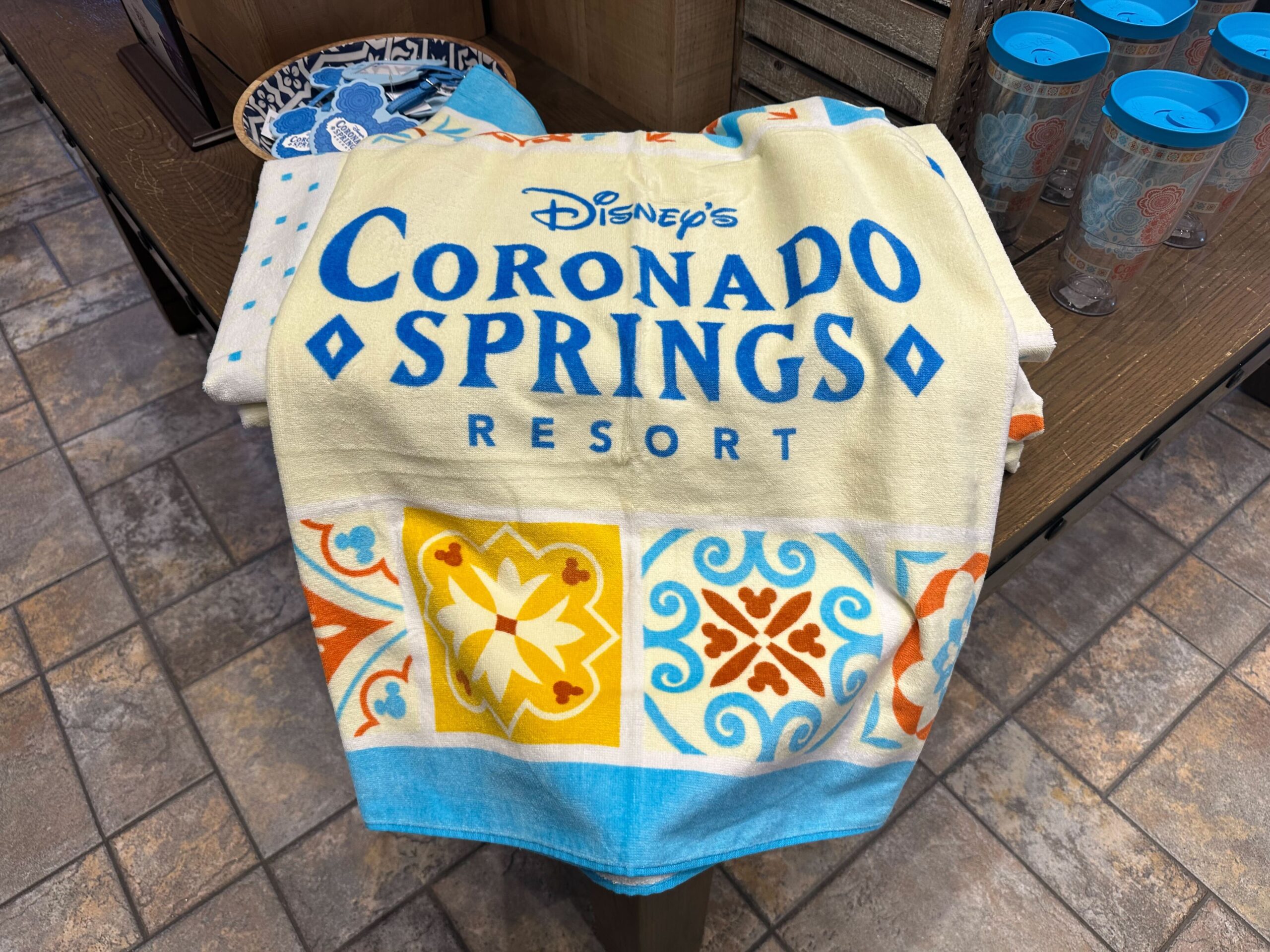 A towel, part of the Coronado Springs merchandise, graces the store shelf with Disney's Coronado Springs Resort text. It showcases vibrant patterns in yellow, orange, and blue hues.'s Coronado Springs Resort text. It showcases vibrant patterns in yellow, orange, and blue hues.