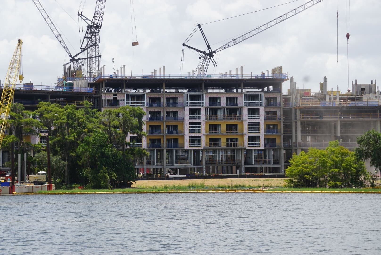 Mid-rise building under construction with cranes, seen across a body of water, and trees in the foreground.