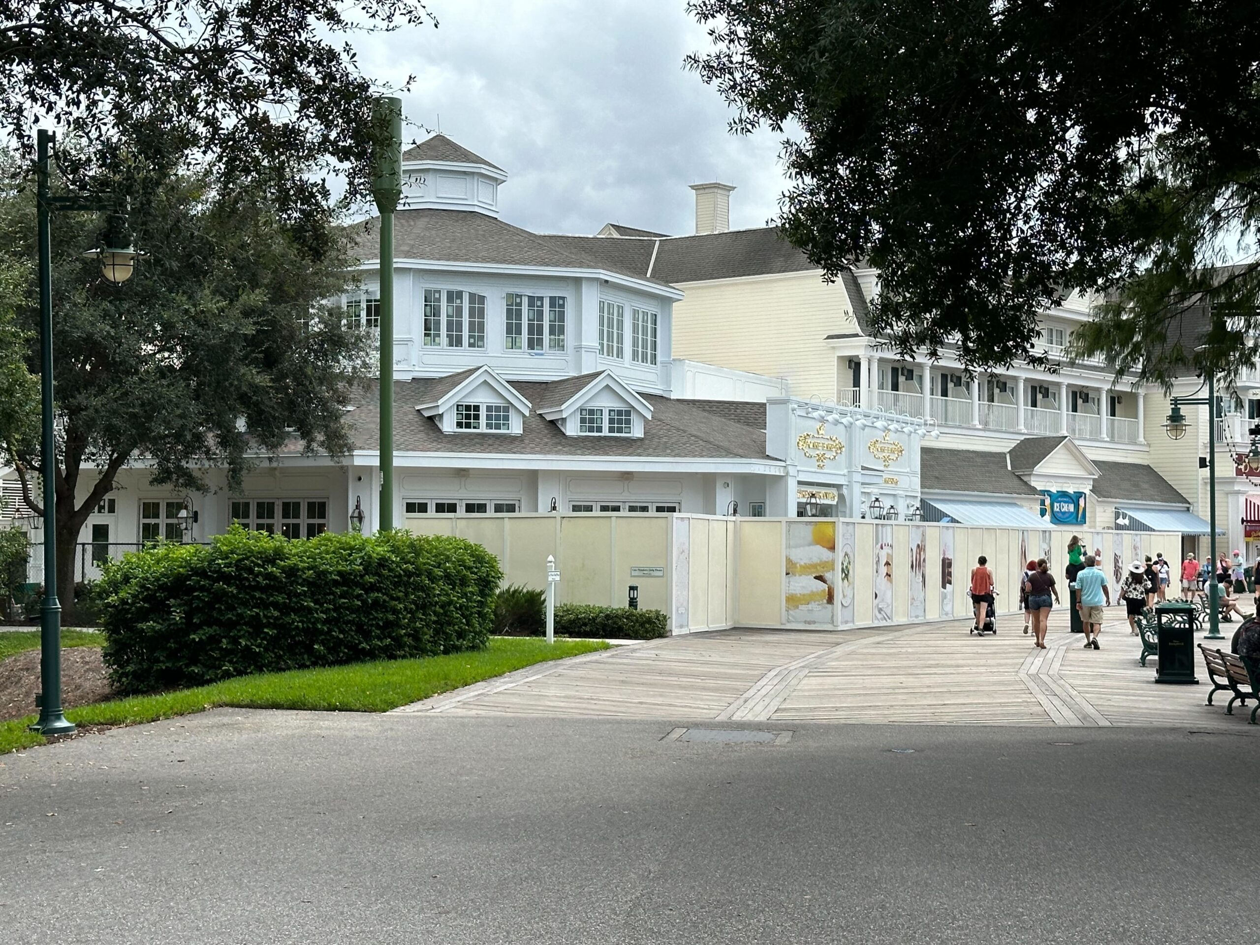 A building with a section under construction, surrounded by trees and a paved walkway. Several people are walking nearby, and the sky is cloudy.