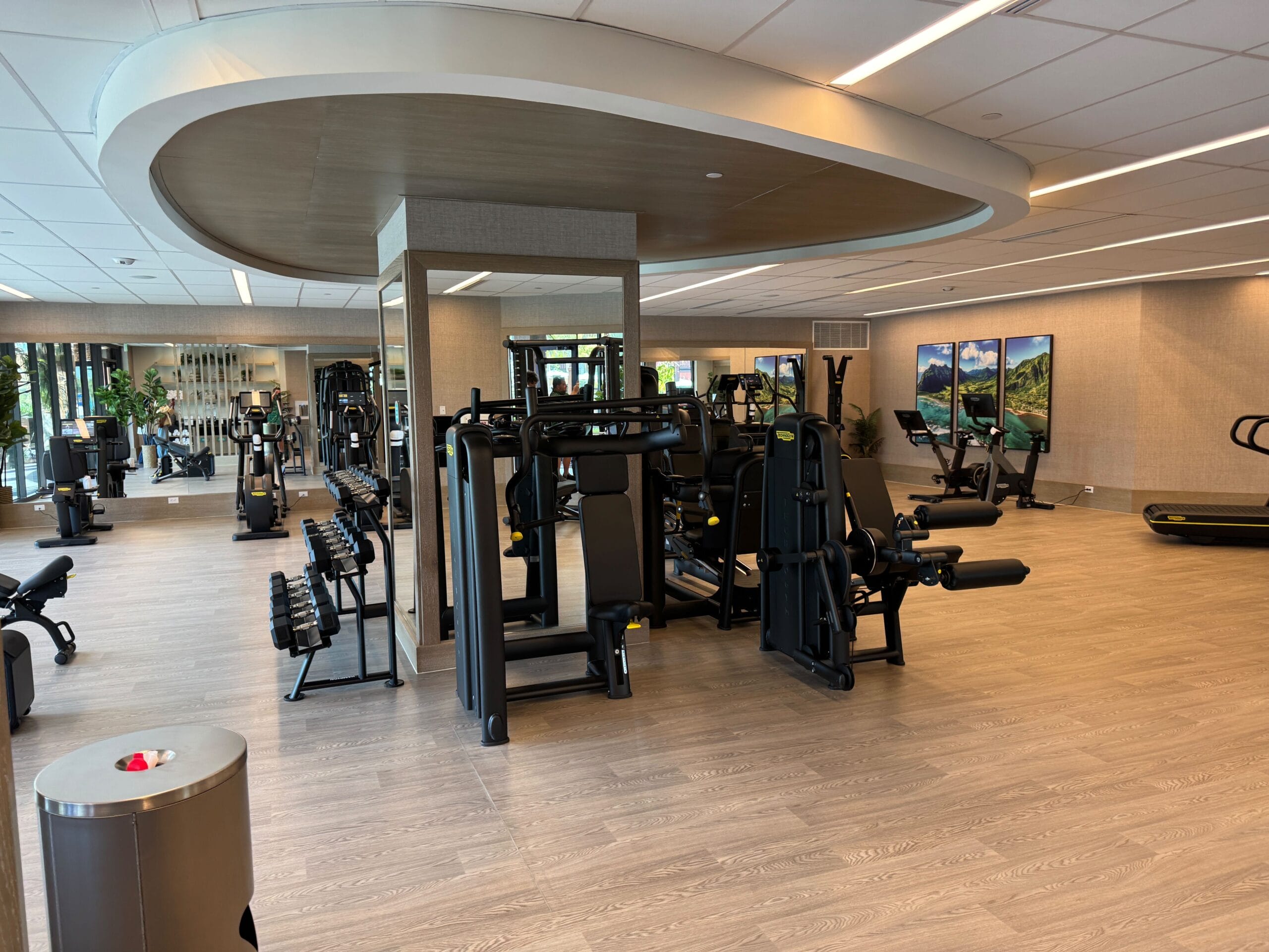 A modern gym with various exercise machines, free weights, and treadmill stations in a spacious room with wooden flooring and large mirrors.