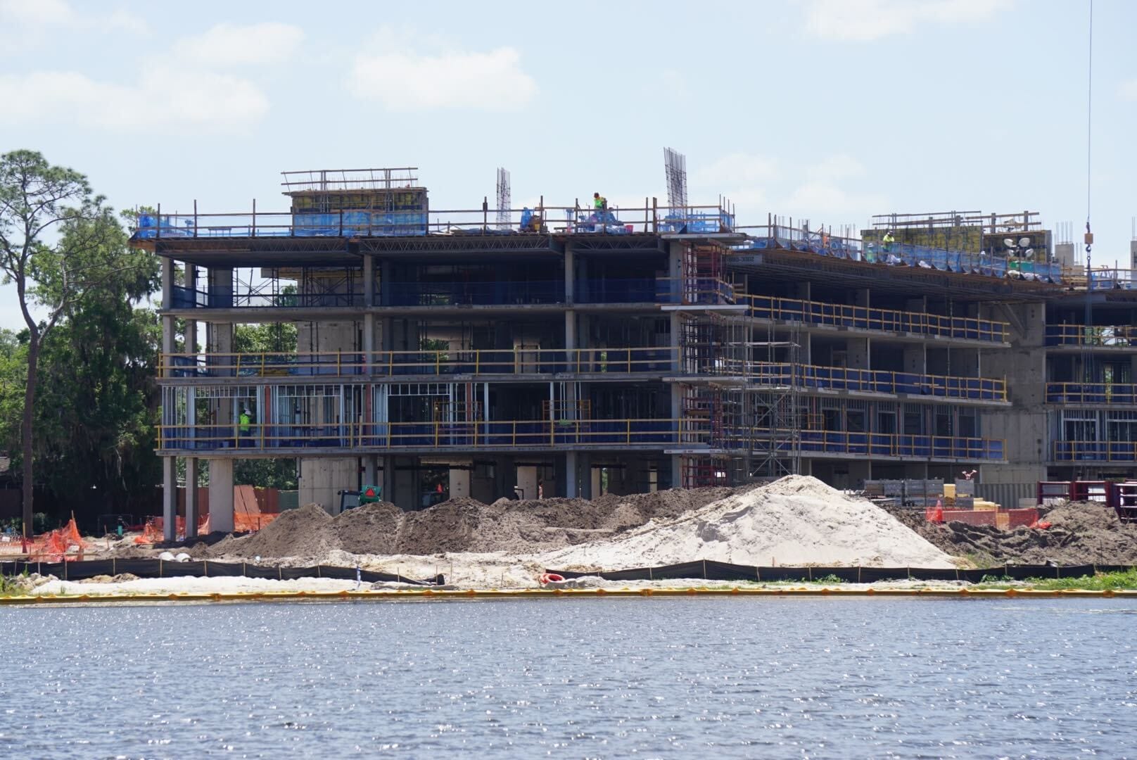 A multi-story building under construction near a body of water, surrounded by scaffolding and construction materials, with several workers on site.