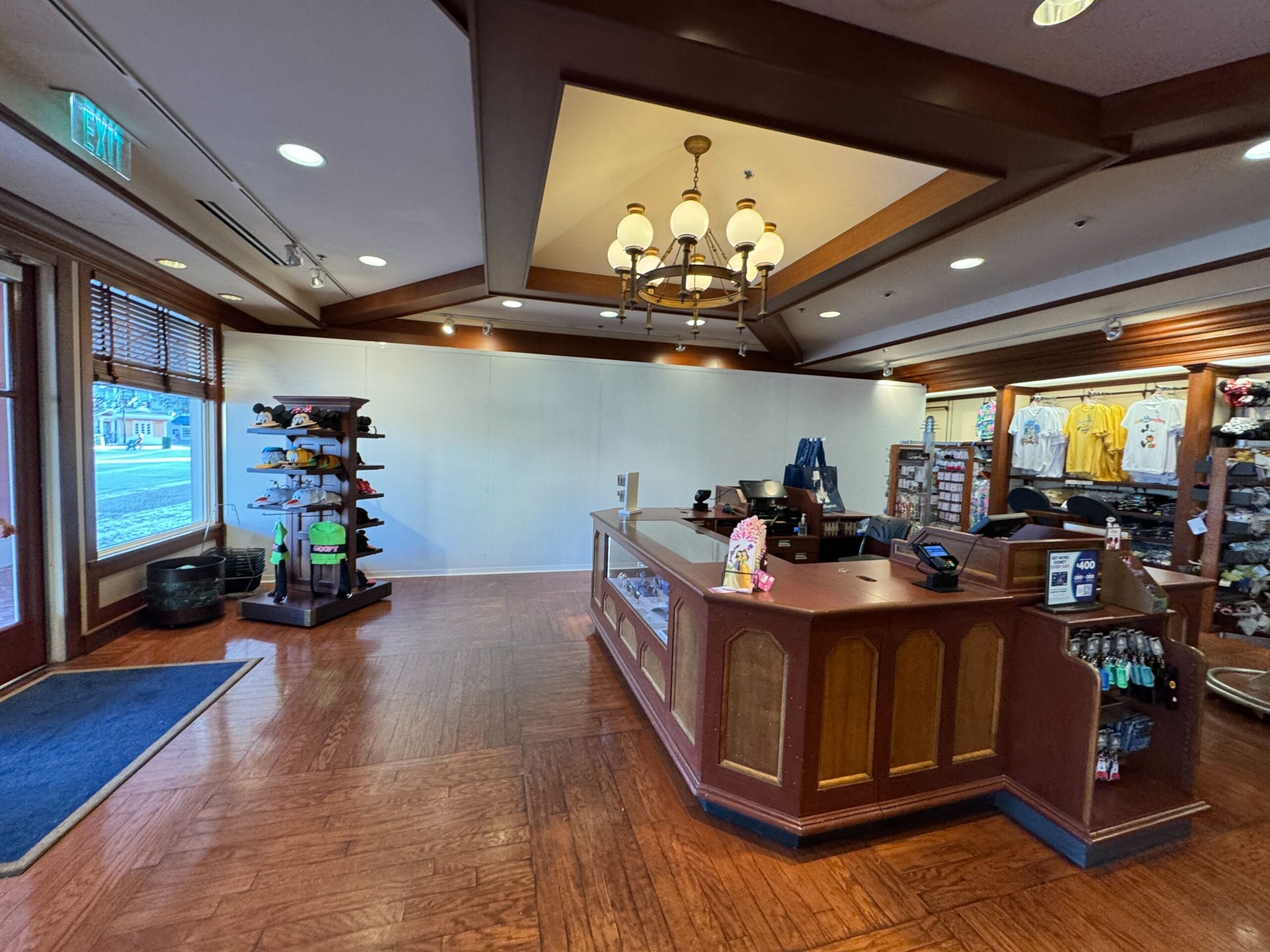 A retail store interior with wooden flooring, a central counter, shelves holding caps and toys, and a wall display of T-shirts. A chandelier hangs from the ceiling.