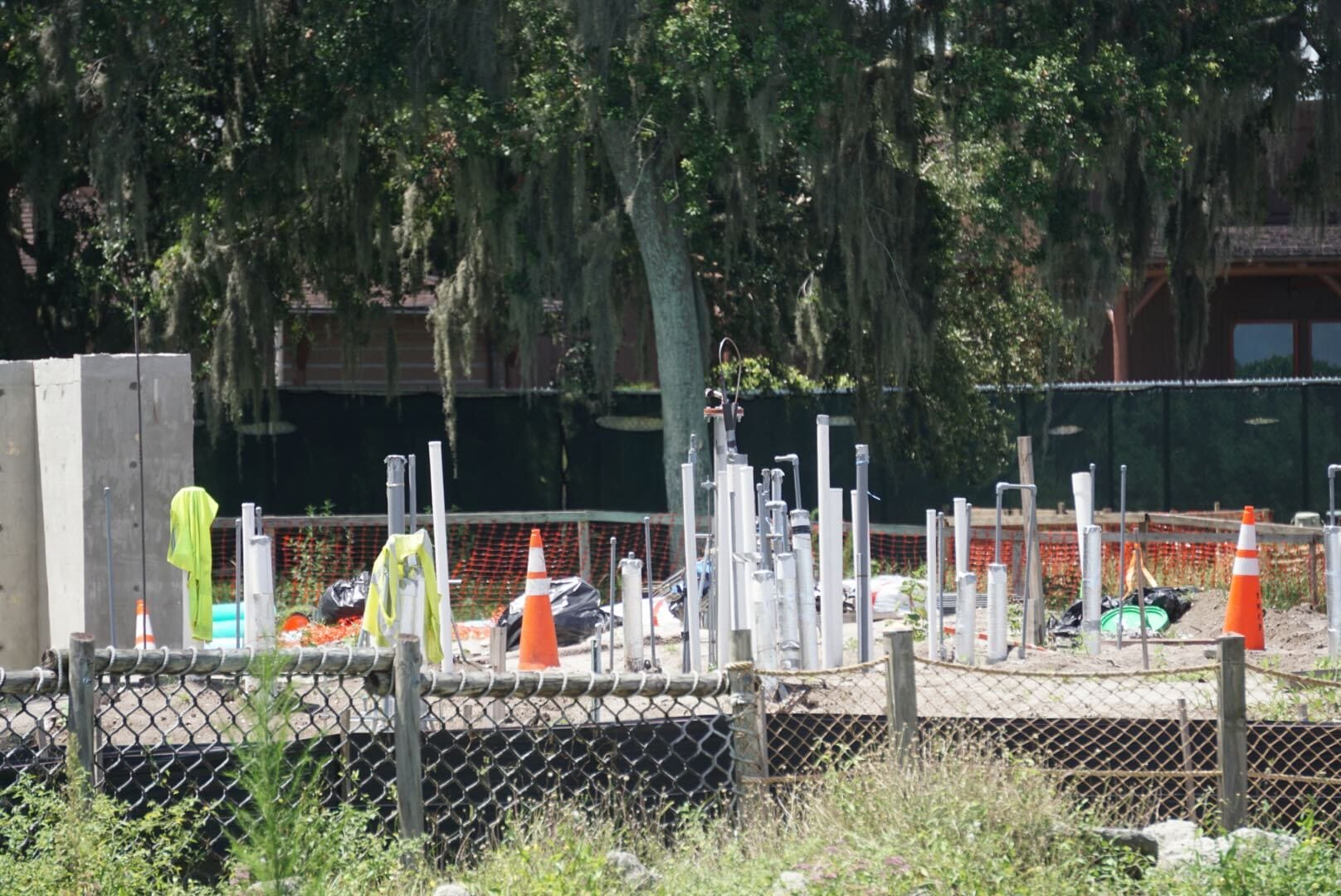 A construction site with pipes, safety cones, fencing, and safety vests, surrounded by trees and greenery.