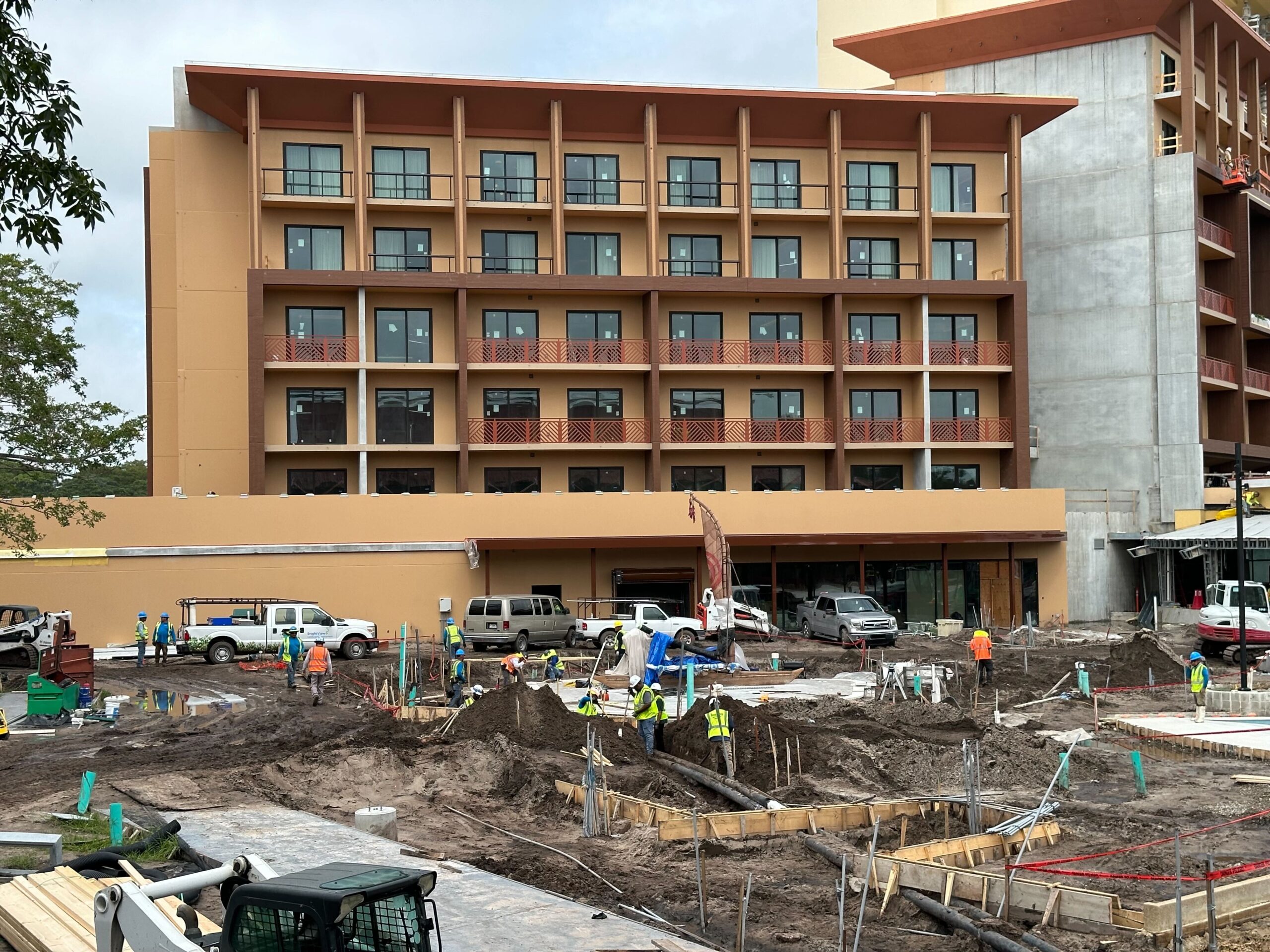 Construction site with workers and equipment in front of a multi-story building under renovation. The site has various materials, vehicles, and scaffolding.